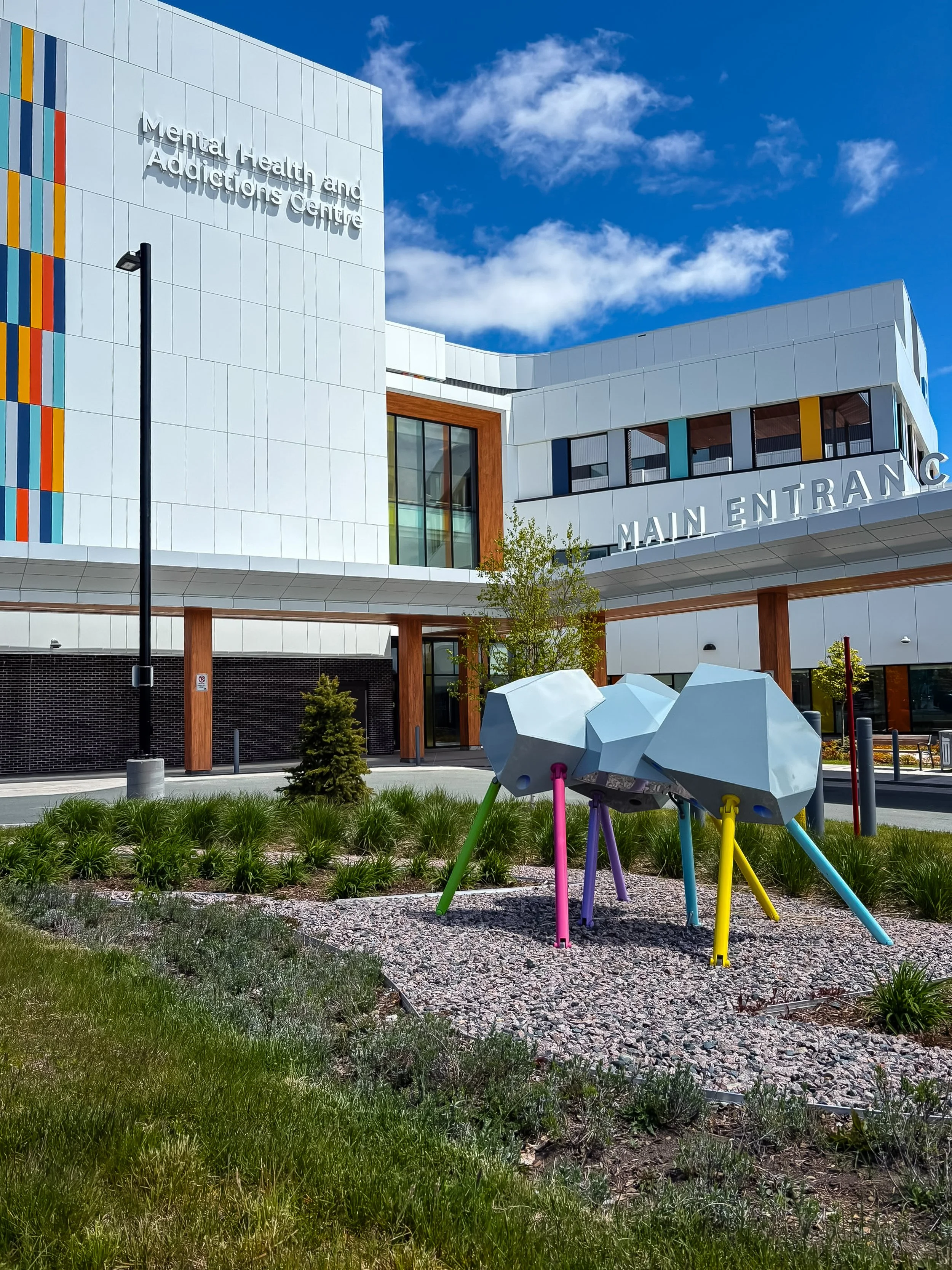 A sculpture of a geometric figure with multi-coloured legs stands outside a large building with "Mental Health and Addictions Centre" written on a wall next to colourful rectangular shapes.