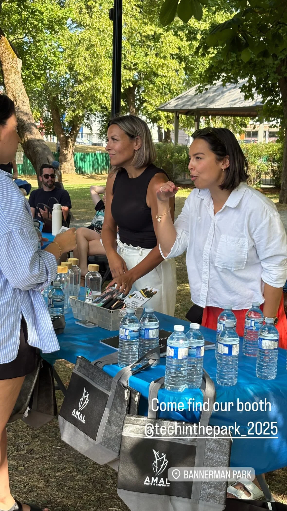 Two women at a table with bottles of water on top speak to another woman standing in front. There are green trees and a gazebo in the background.