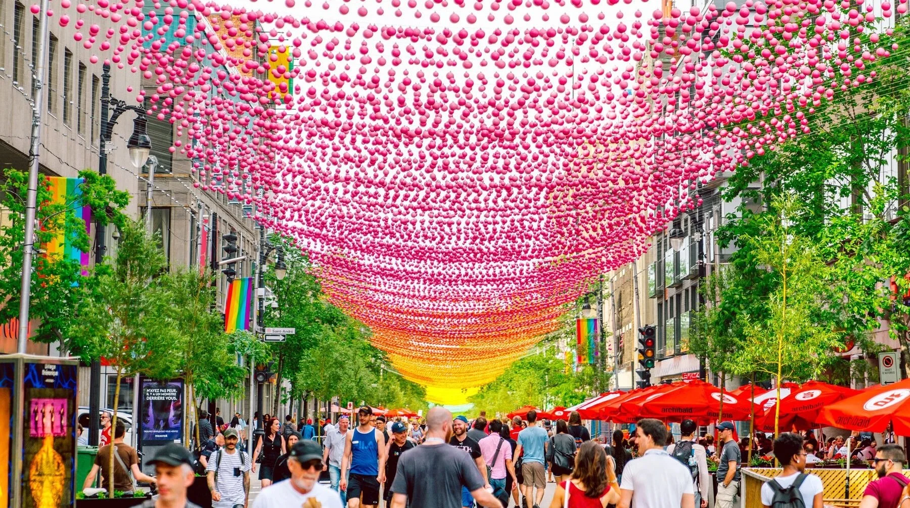 A large swath of pink circular shapes hangs over a street, with rainbow-colured flags hung outside several buildings, as groups of people walk underneath.