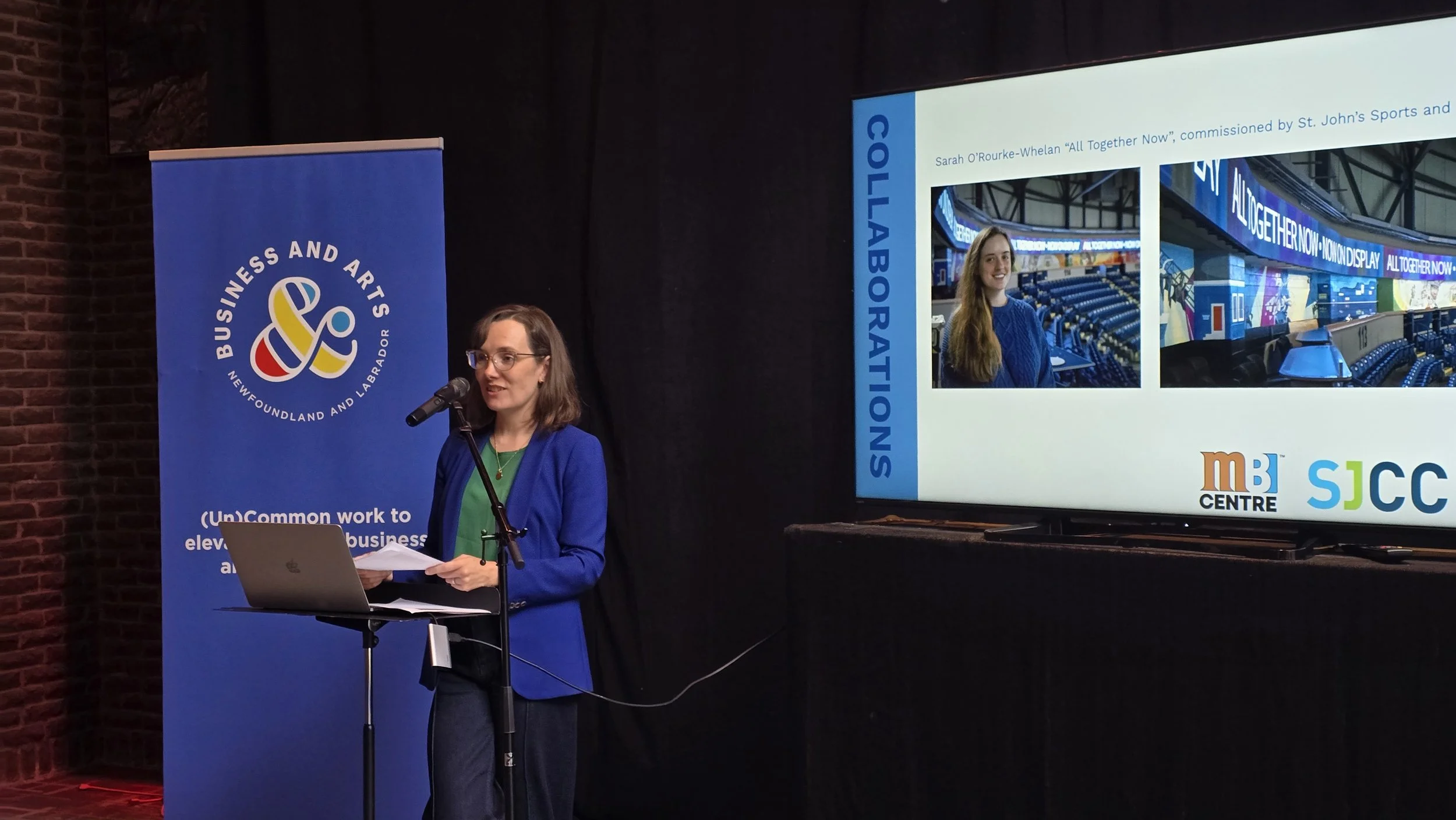 A woman in a blue blazer, green shirt and glasses with shoulder length dark hair speaks into a mic at a podium. Behind her is a blue Business & Arts NL banner and a screen showing a photo of a smiling woman next to a photo of a mural.