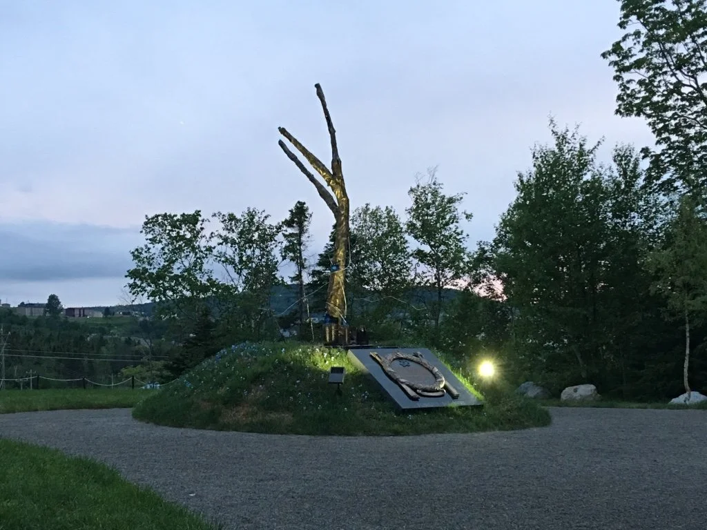 A sculpture of a tree branch with a plaque underneath juts out of a small green hill with trees around it.