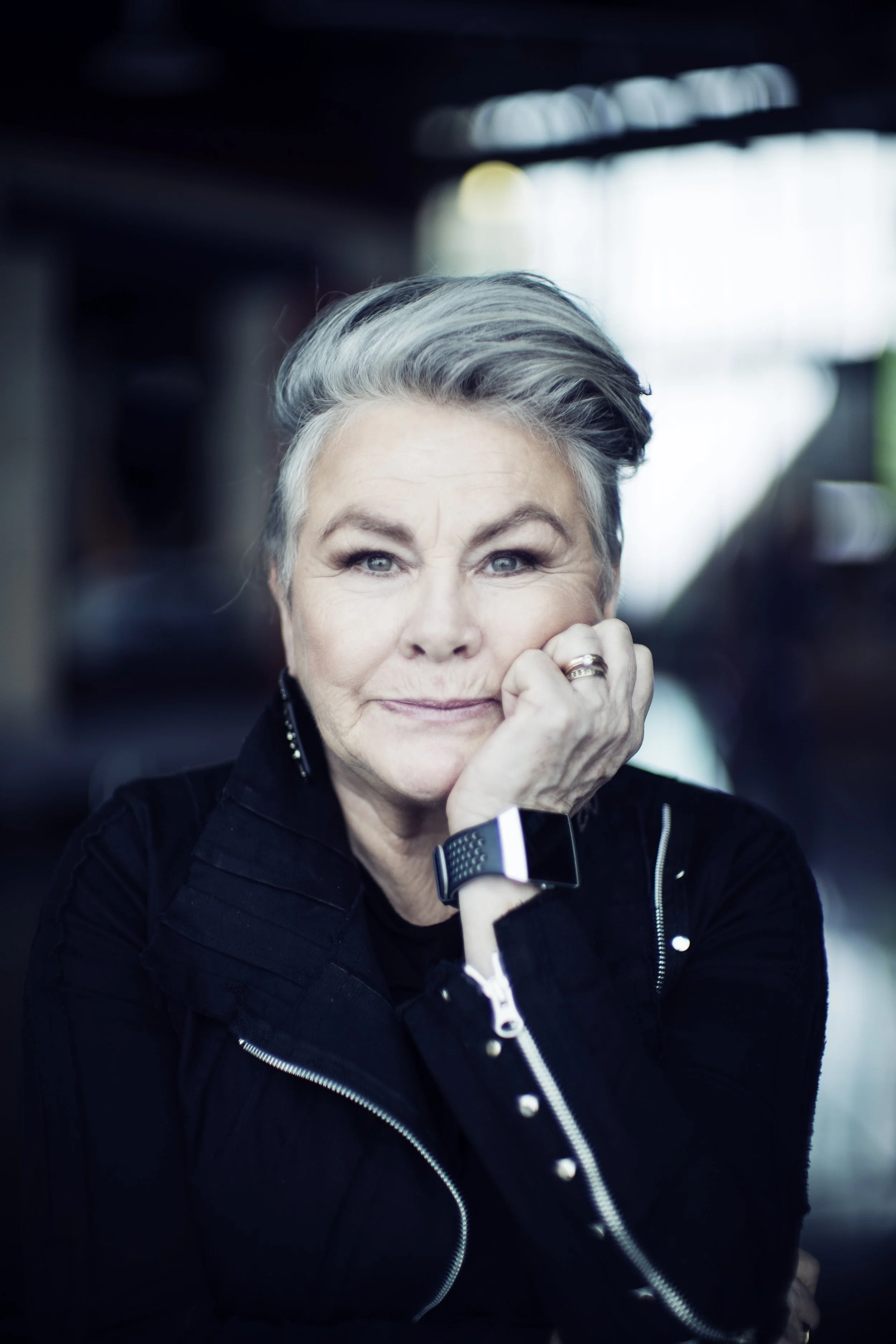 A woman with short, grey hair, wearing a zippered jacket, looks at the camera  while holding her left hand under her chin.