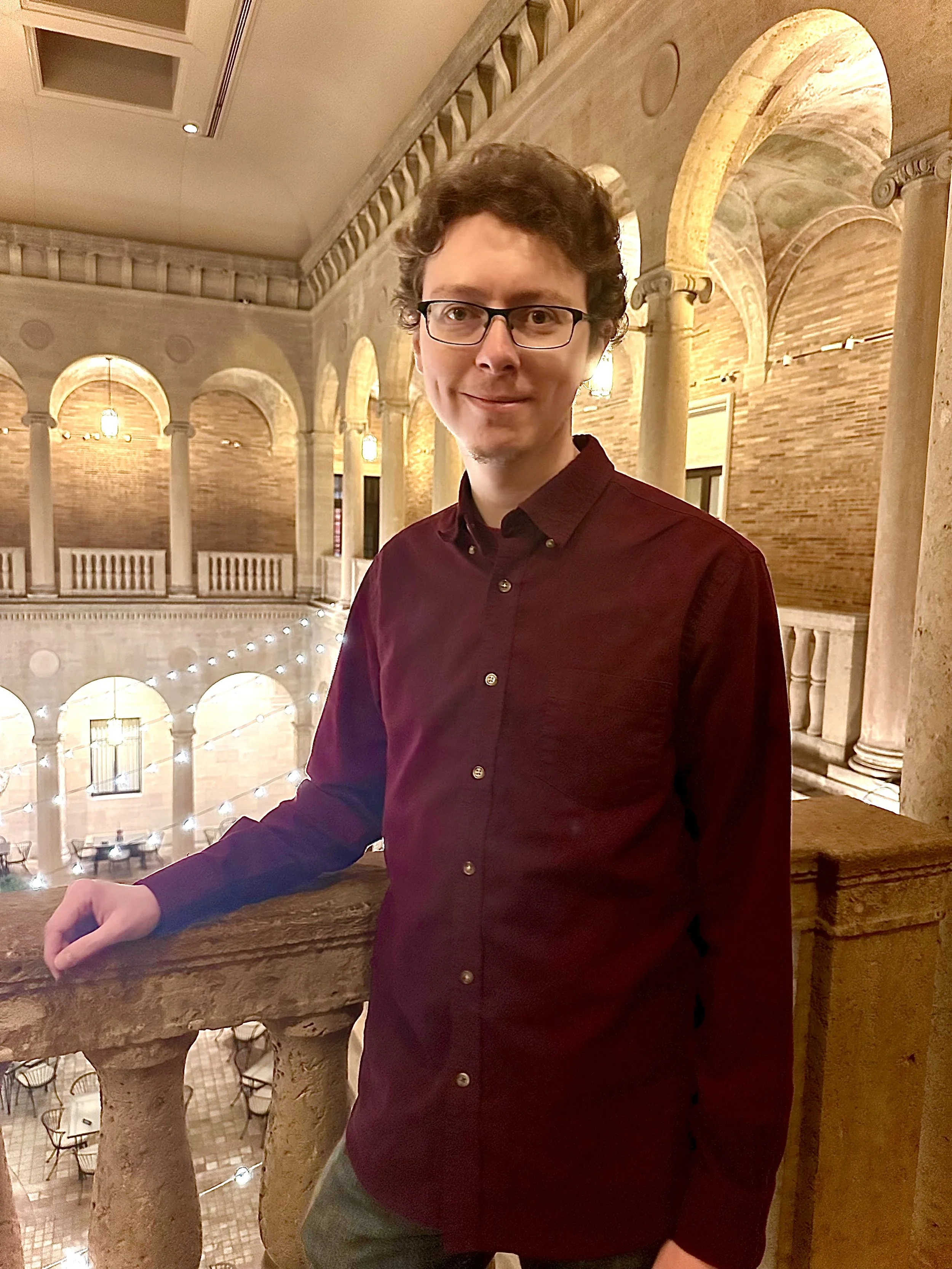 A young man with glasses, curly brown hair, and a slight smile, wearing a burgundy button-up shirt, standing indoors near a stone railing in a building with arched columns, brick walls, and warm lighting.