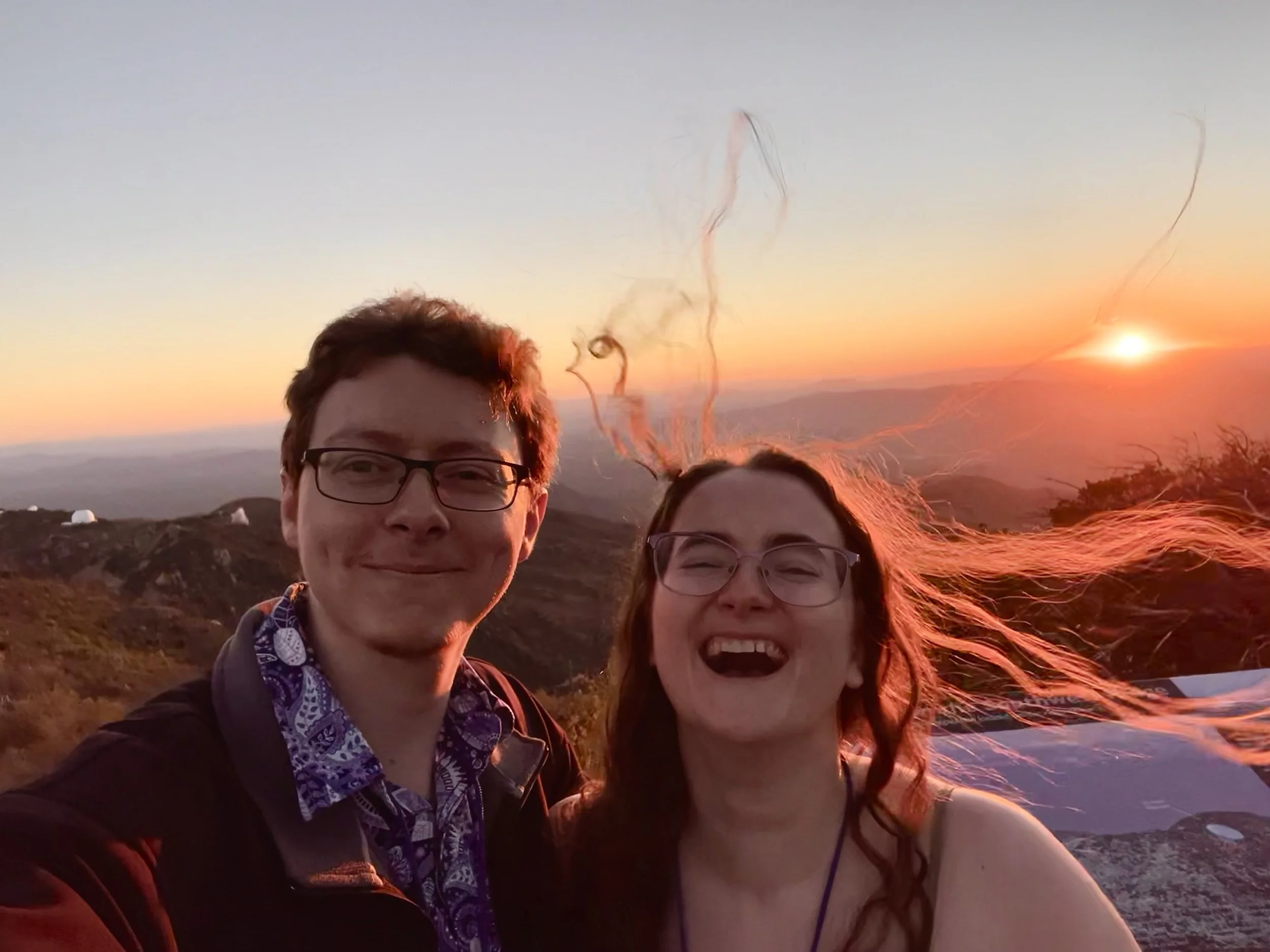 Two smiling people, a man and a woman, taking a selfie during sunset with a mountainous landscape in the background.