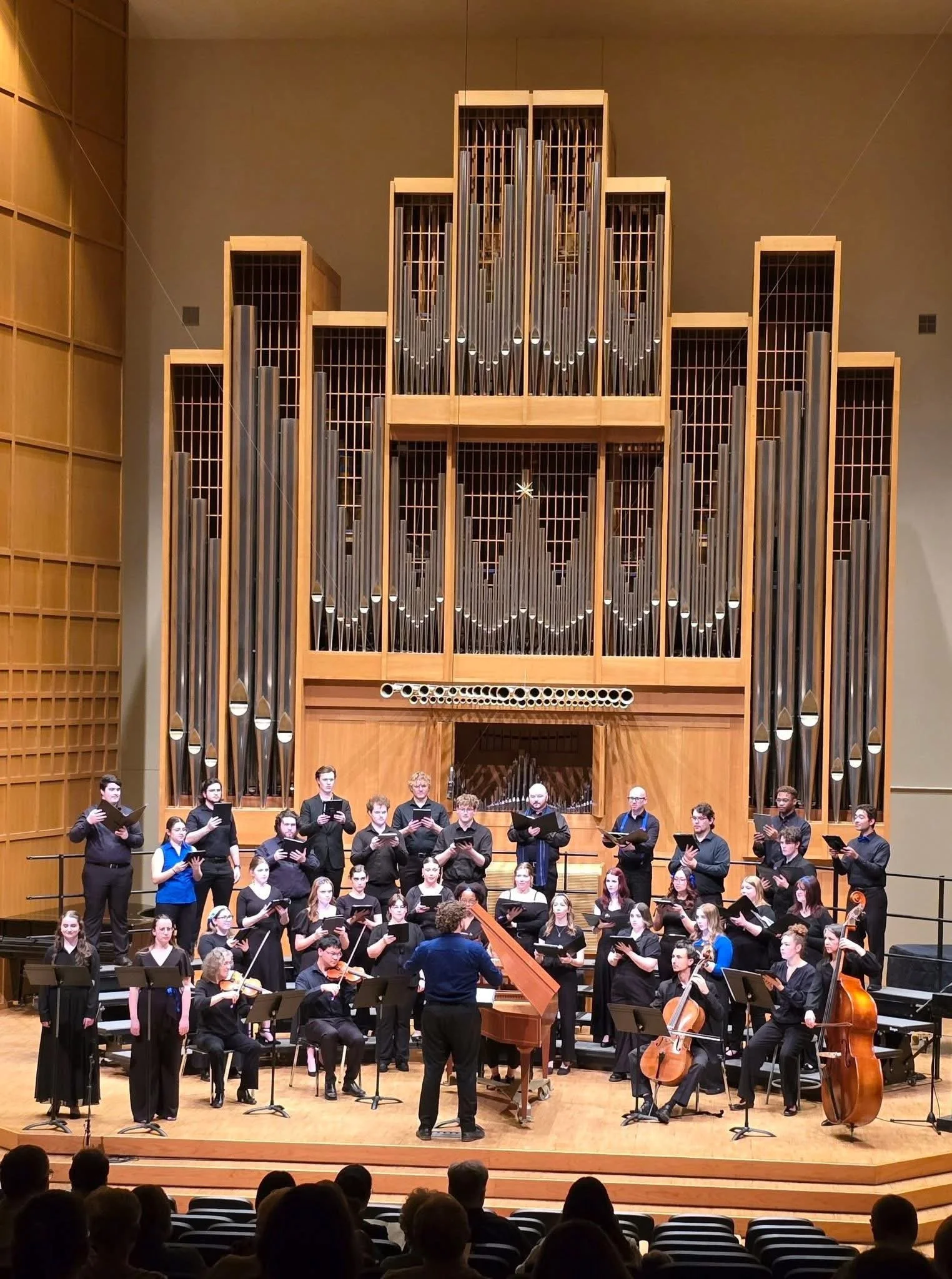 A large choir group performing on stage with a conductor and an orchestra, high organ pipes in the background, in a concert hall.