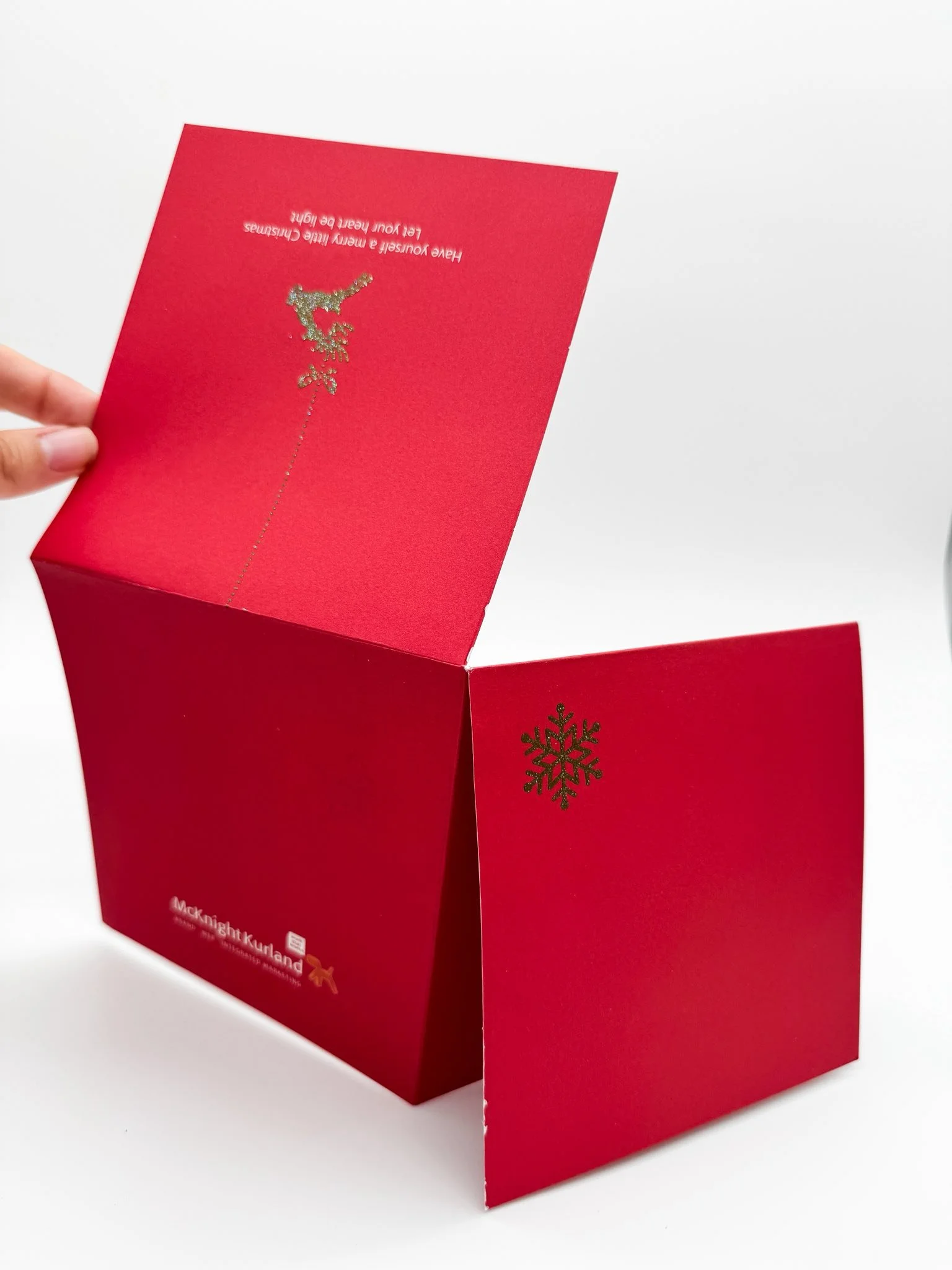 Red Square-shaped Christmas card with gold glitter snowflake and custom scored folds being held open by a hand, with white background.