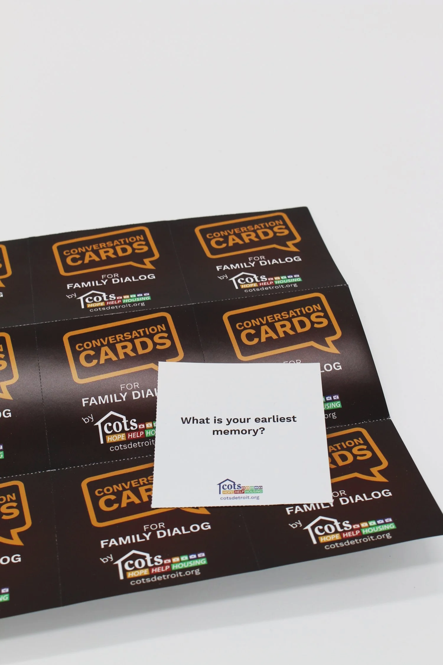 Conversation cards for family dialogue by COTS, with a question on a white card asking, 'What is your earliest memory?' placed on a black background with multiple cards.