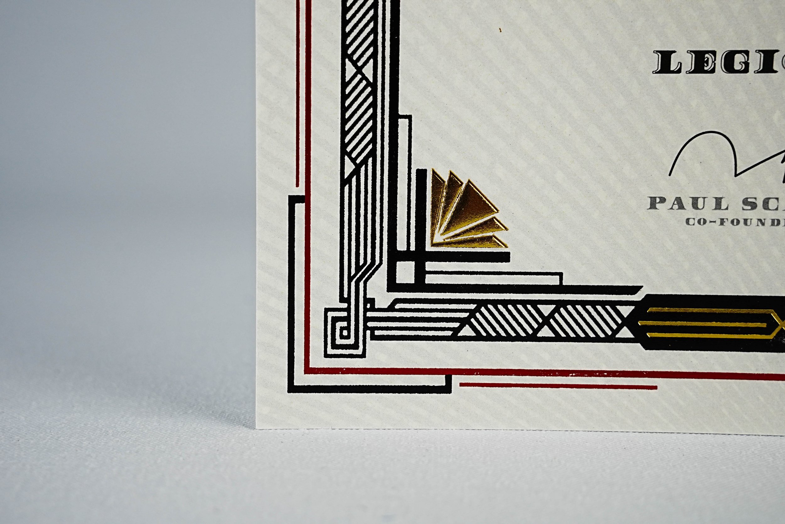 Close-up of an ornate certificate corner design featuring black, red, and gold metallic foil geometric patterns with partial text and signature.