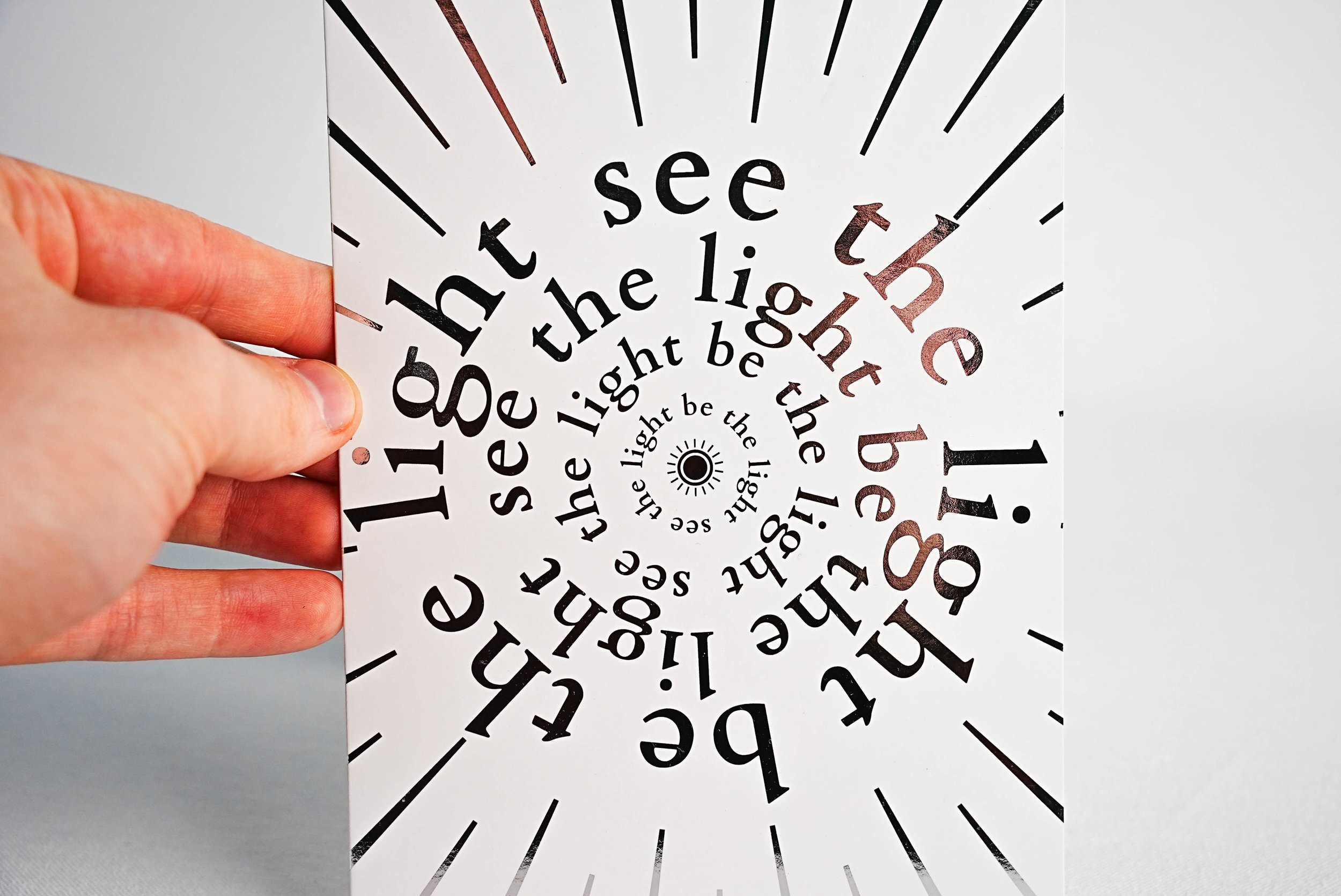 Hand holding a card with spiraling text "see the light" and "be the light" that was printed by TEAM Concept using metallic foil printing