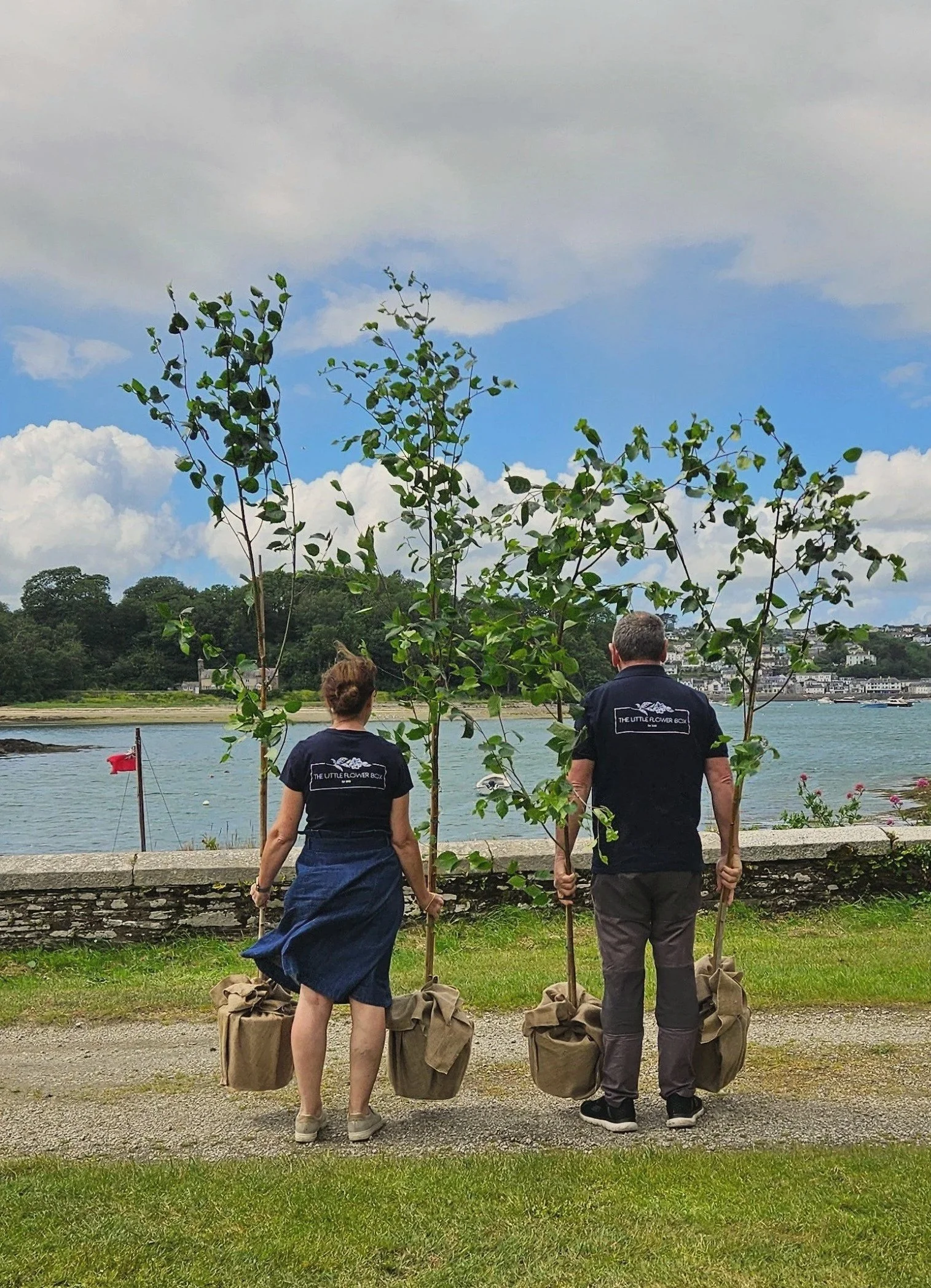 Two people standing outdoors near water, holding saplings in burlap bags, with trees and a waterfront city in the background, wearing matching shirts that say 'The Little Flower Box'.
