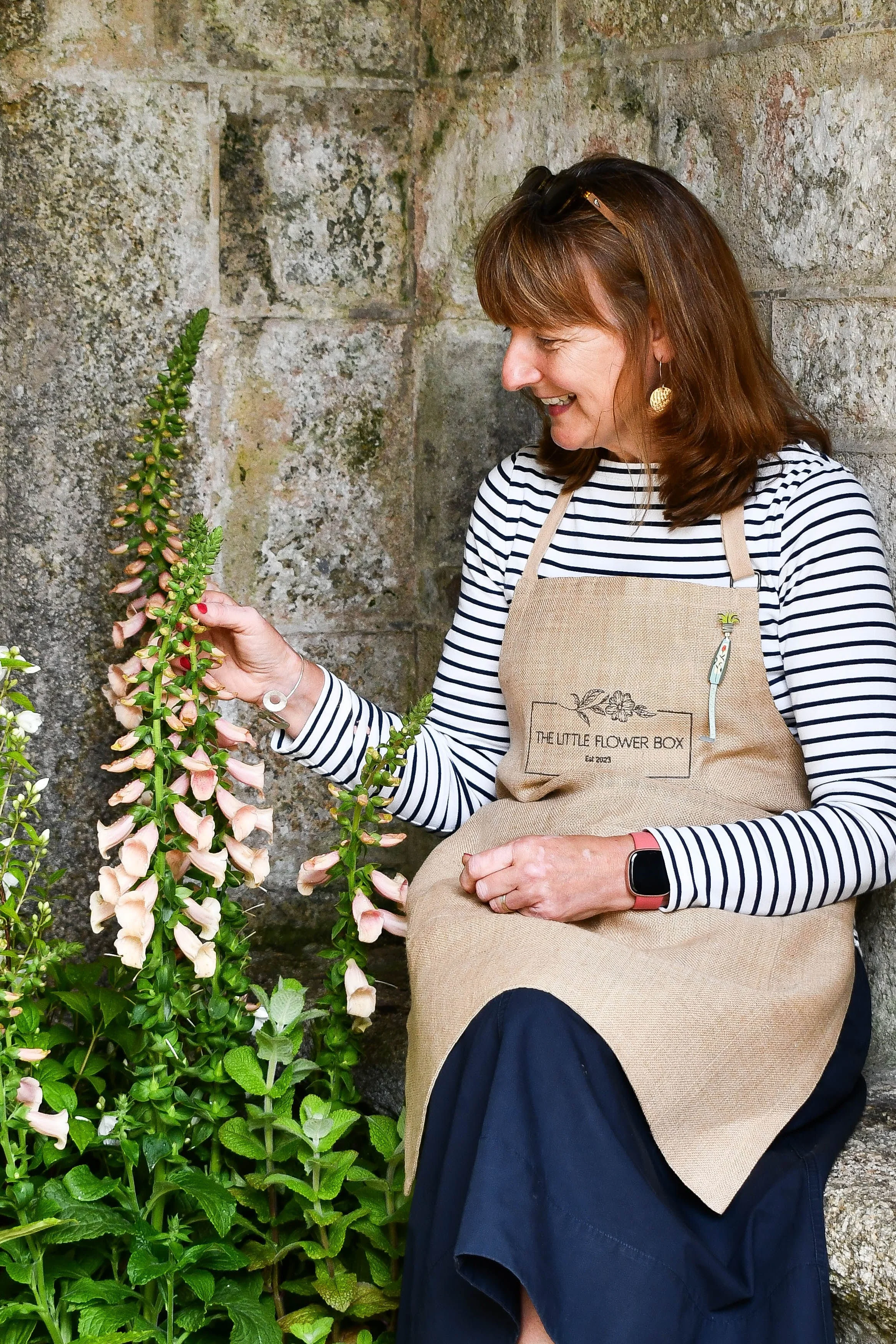 A woman wearing a striped shirt, beige apron, and navy skirt is sitting against a stone wall, smiling while holding a stem of pink and cream flowers.