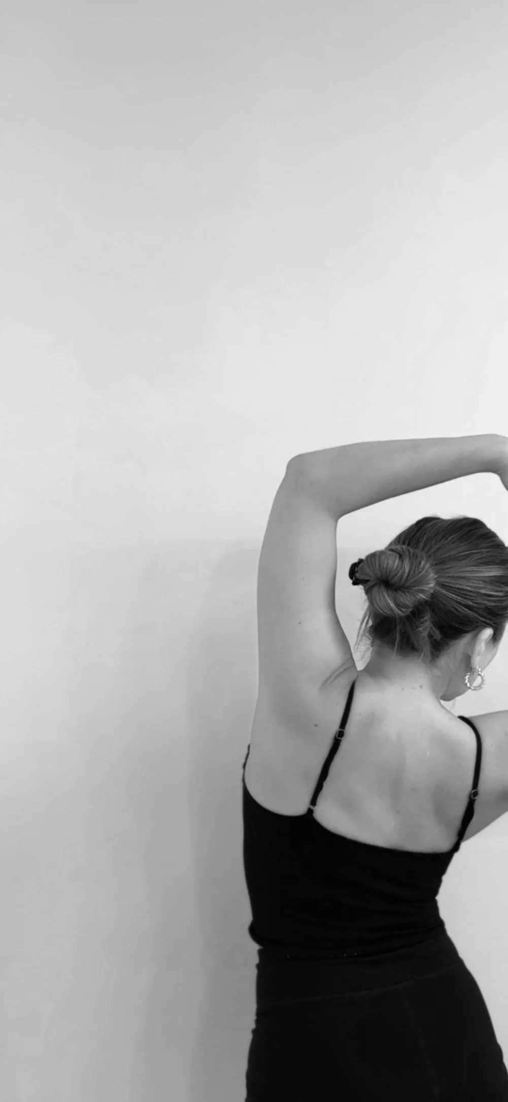 Black and white image of a woman stretching with her back turned, wearing a tank top, hair in a bun.