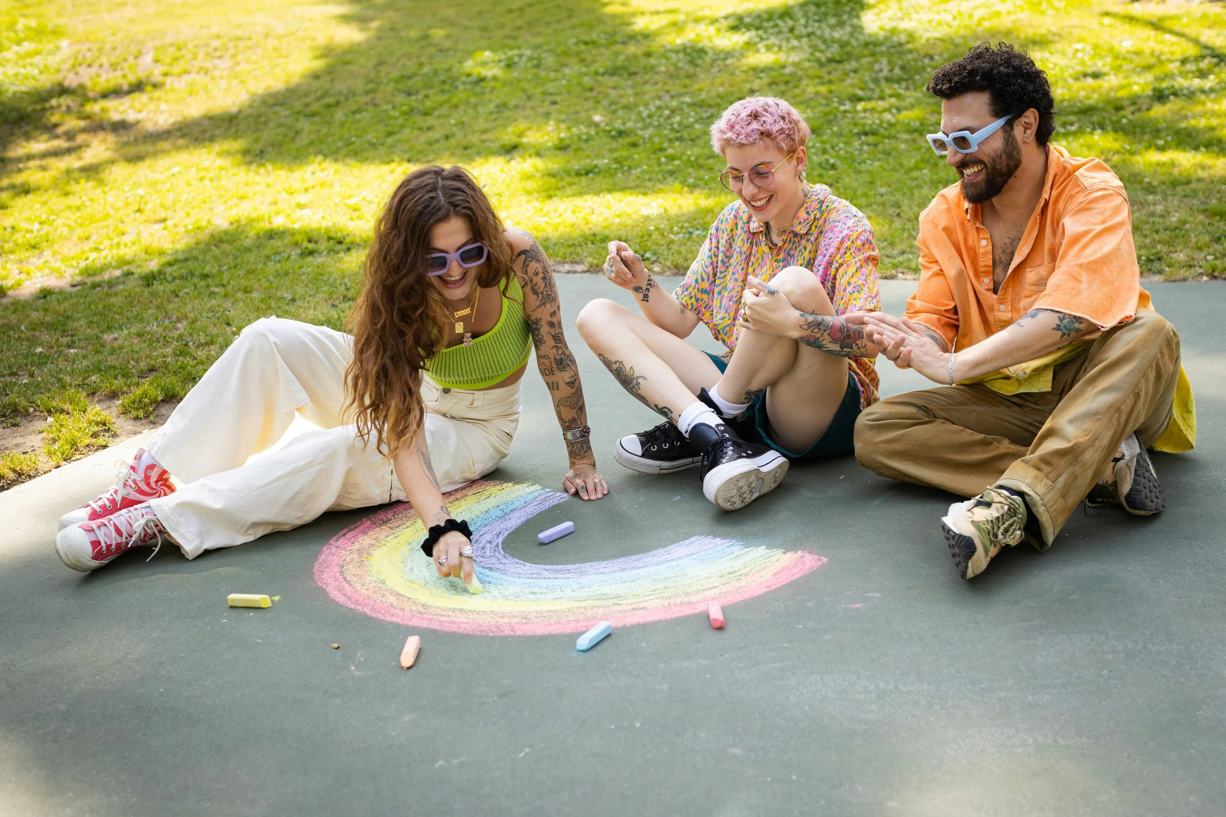 Three adults sit on the pavement and draw a rainbow with chalk
