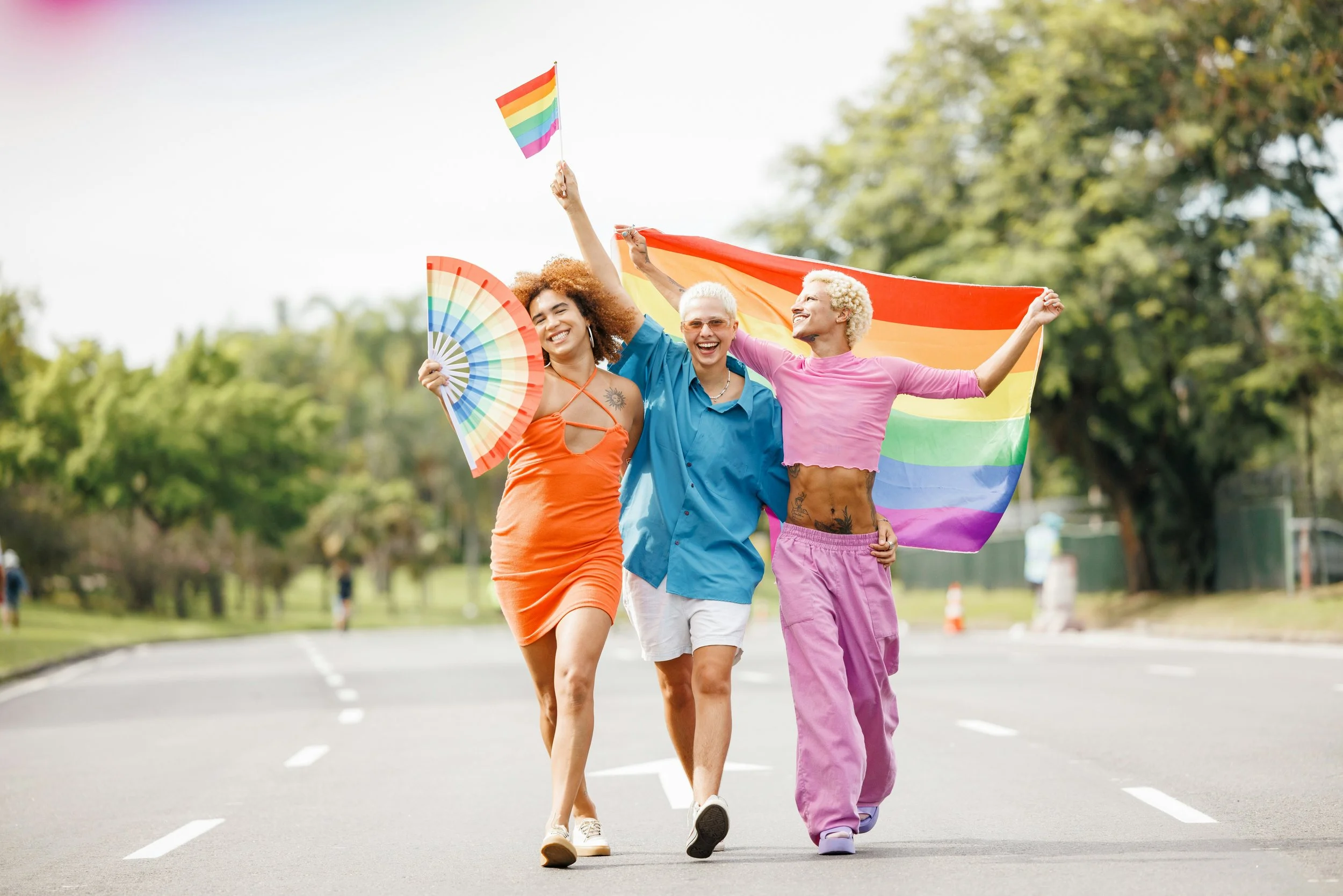 Three queer adults joyfully walk down the street with pride flags