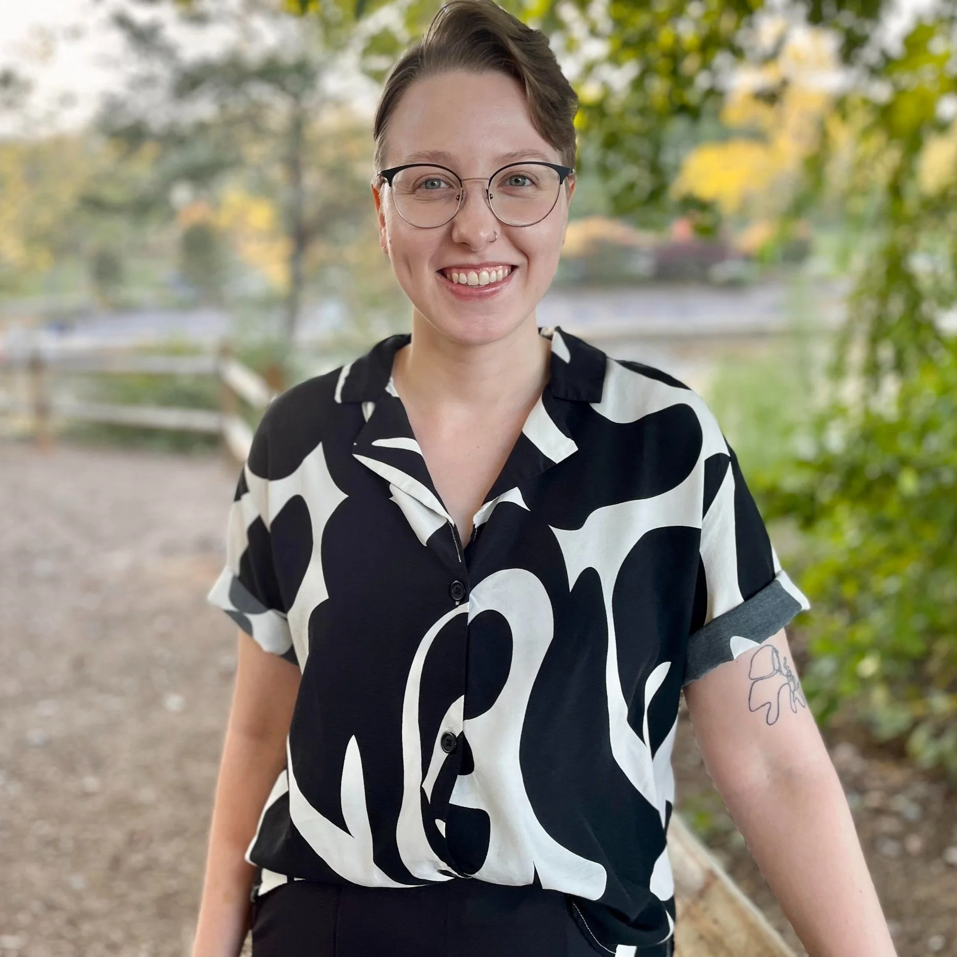Jess, the founder of Colors of You Counseling & Consulting, is pictured with short hair, glasses, a nose ring, and a black and white patterned shirt. They are standing outdoors on a sunny day and smiling happily.