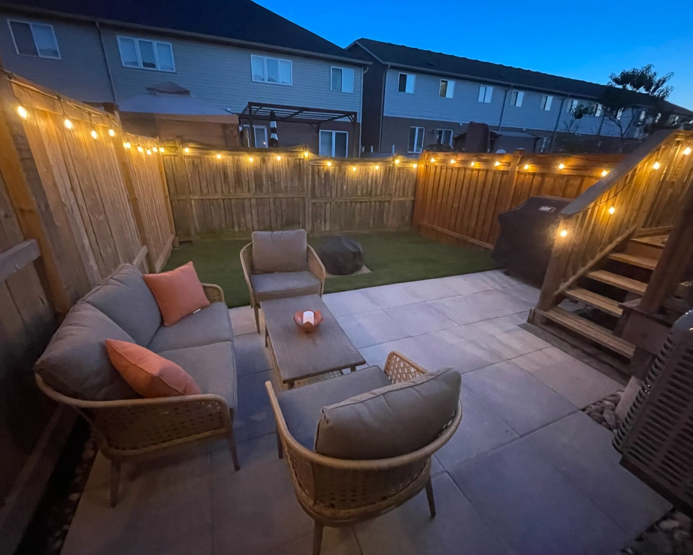 Cozy backyard patio with wicker furniture, string lights, and a grill, surrounded by a wooden fence and neighboring houses.