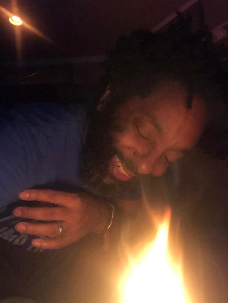 A man with a beard and dreadlocks, wearing a blue shirt and rings, sitting near a fire in a dark environment, laughing and appearing happy.