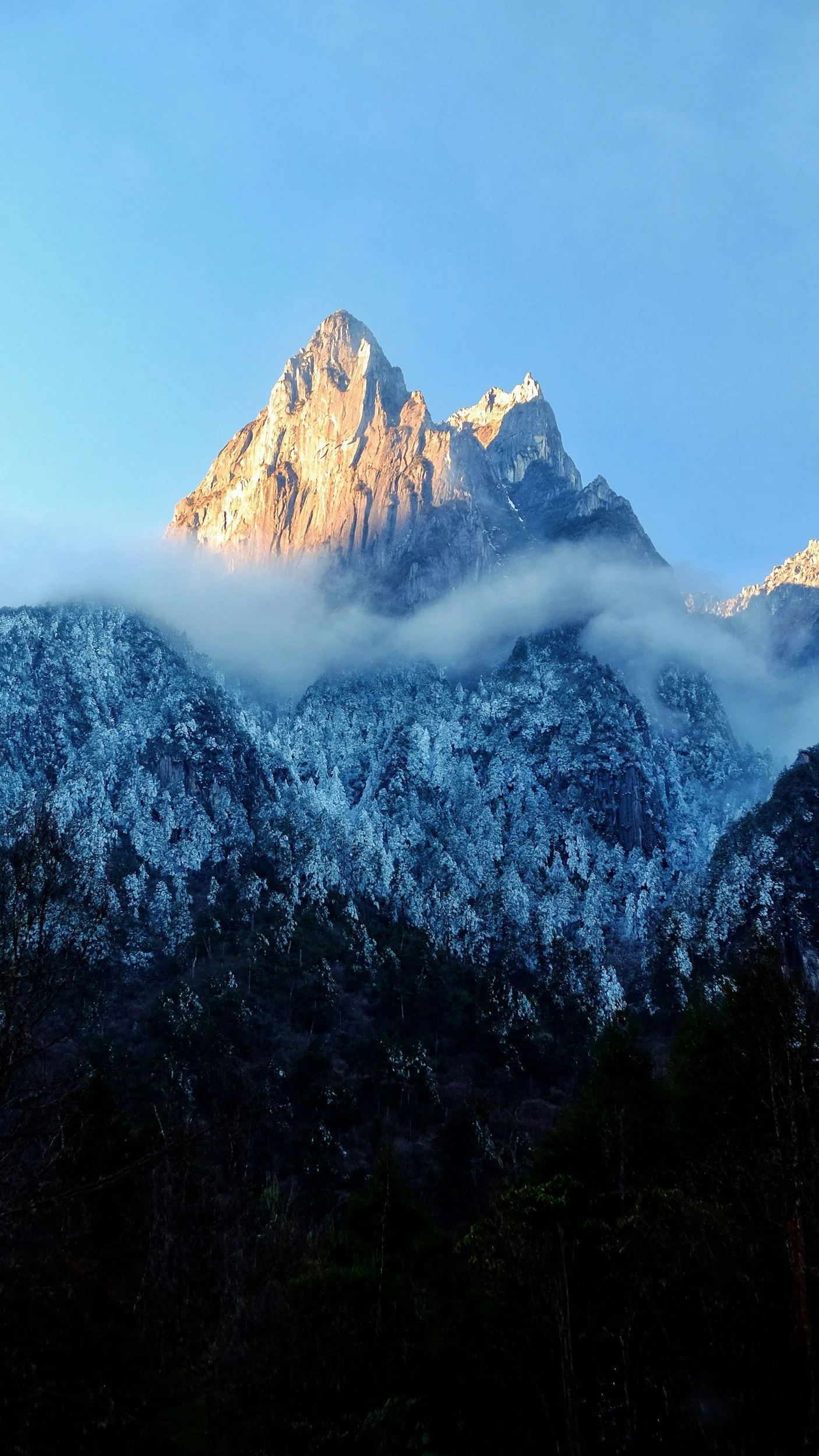 Snow-covered mountain peaks with a prominent rugged summit, partially obscured by a layer of mist, against a clear blue sky.