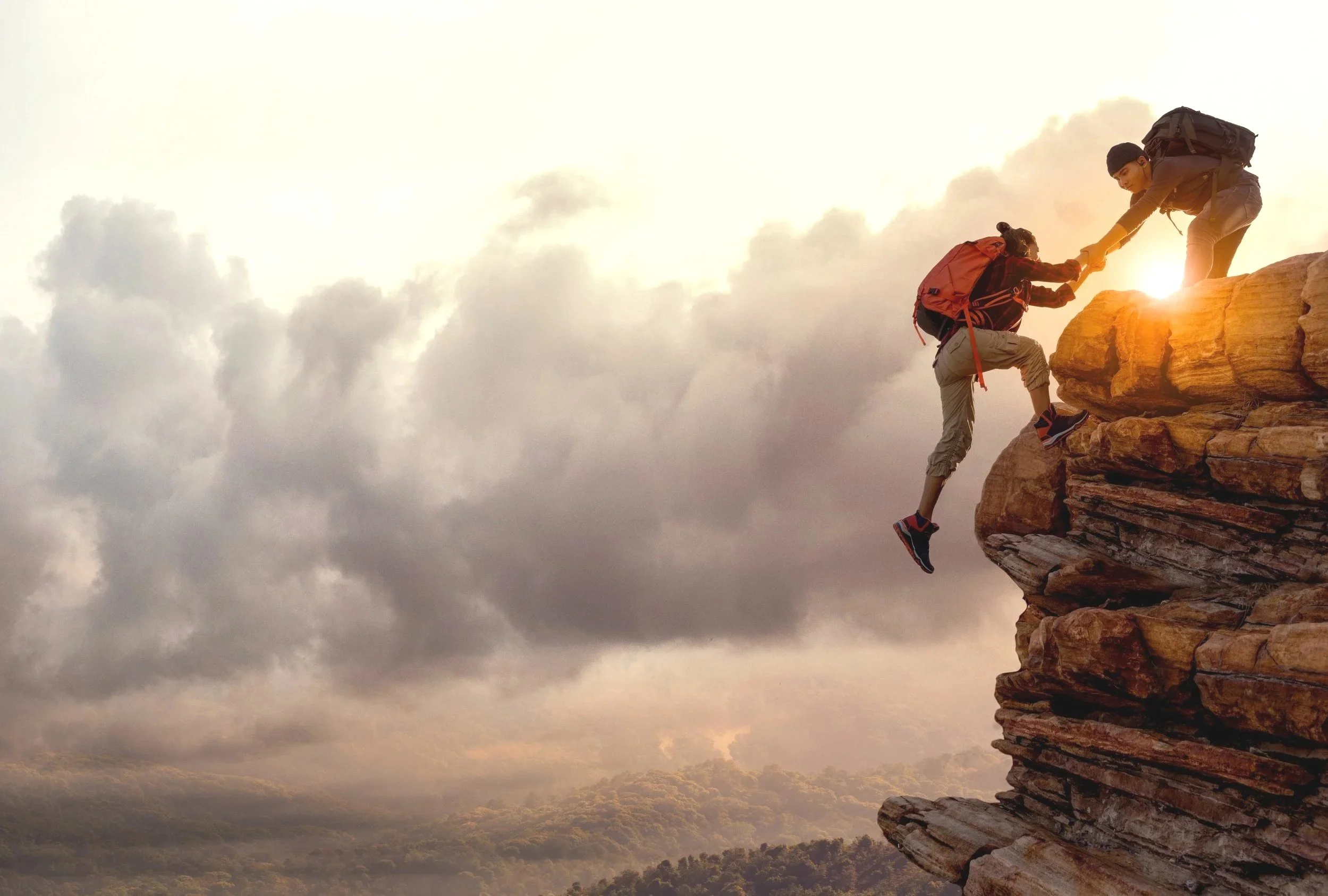 Two hikers on a rocky cliff, one helping the other climb up, with cloudy sky and distant landscape in the background during sunset.