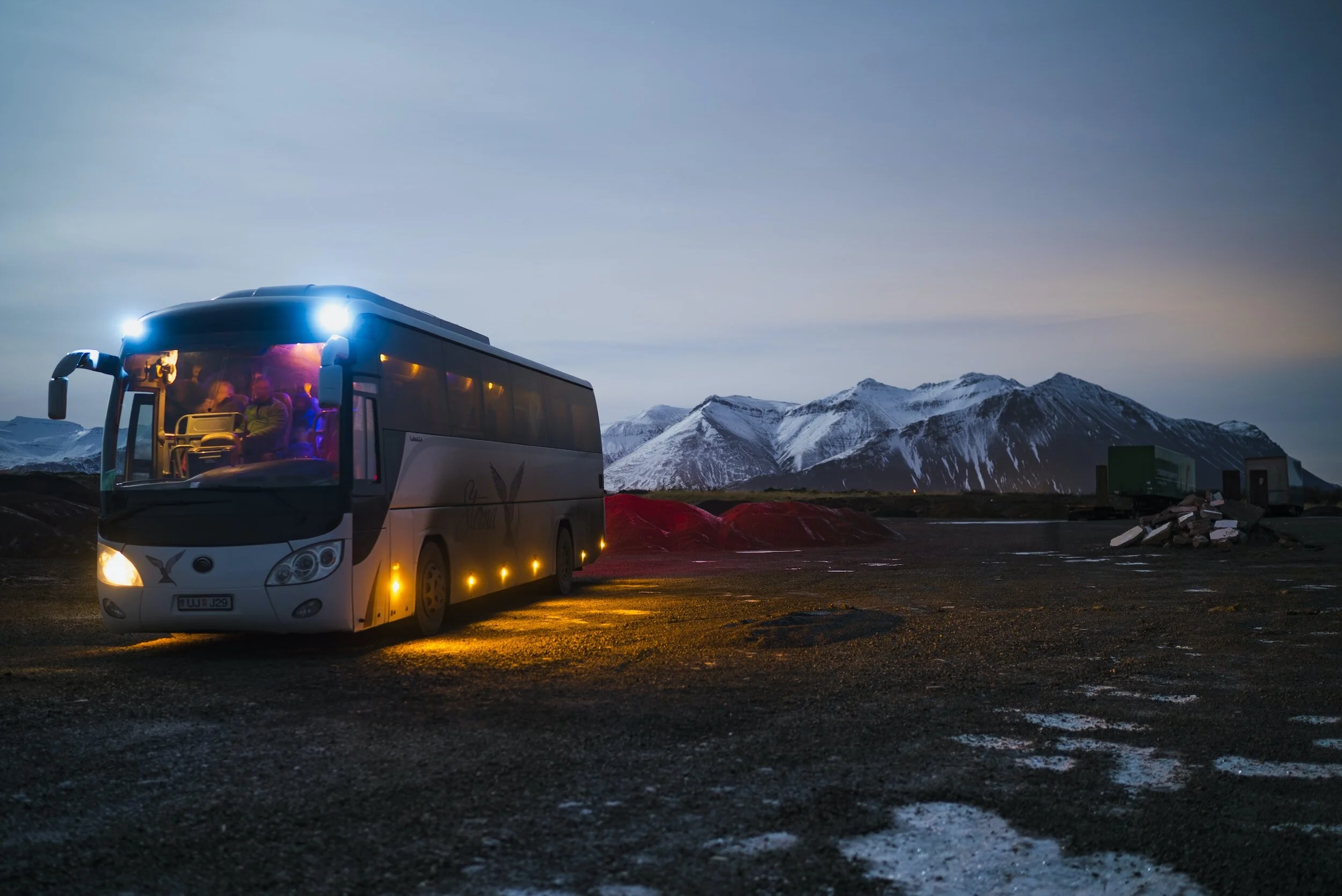 A tour bus parked in a mountainous landscape during twilight with snow-capped peaks in the background and some red and green containers nearby.