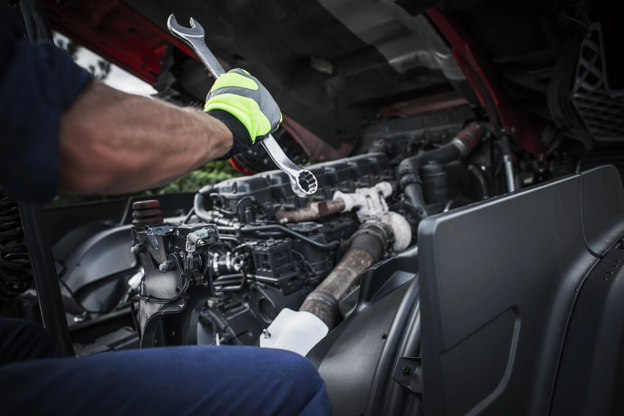 A mechanic working on a truck engine with a large wrench in hand, wearing a black glove with neon yellow accents, under the open hood of the vehicle.