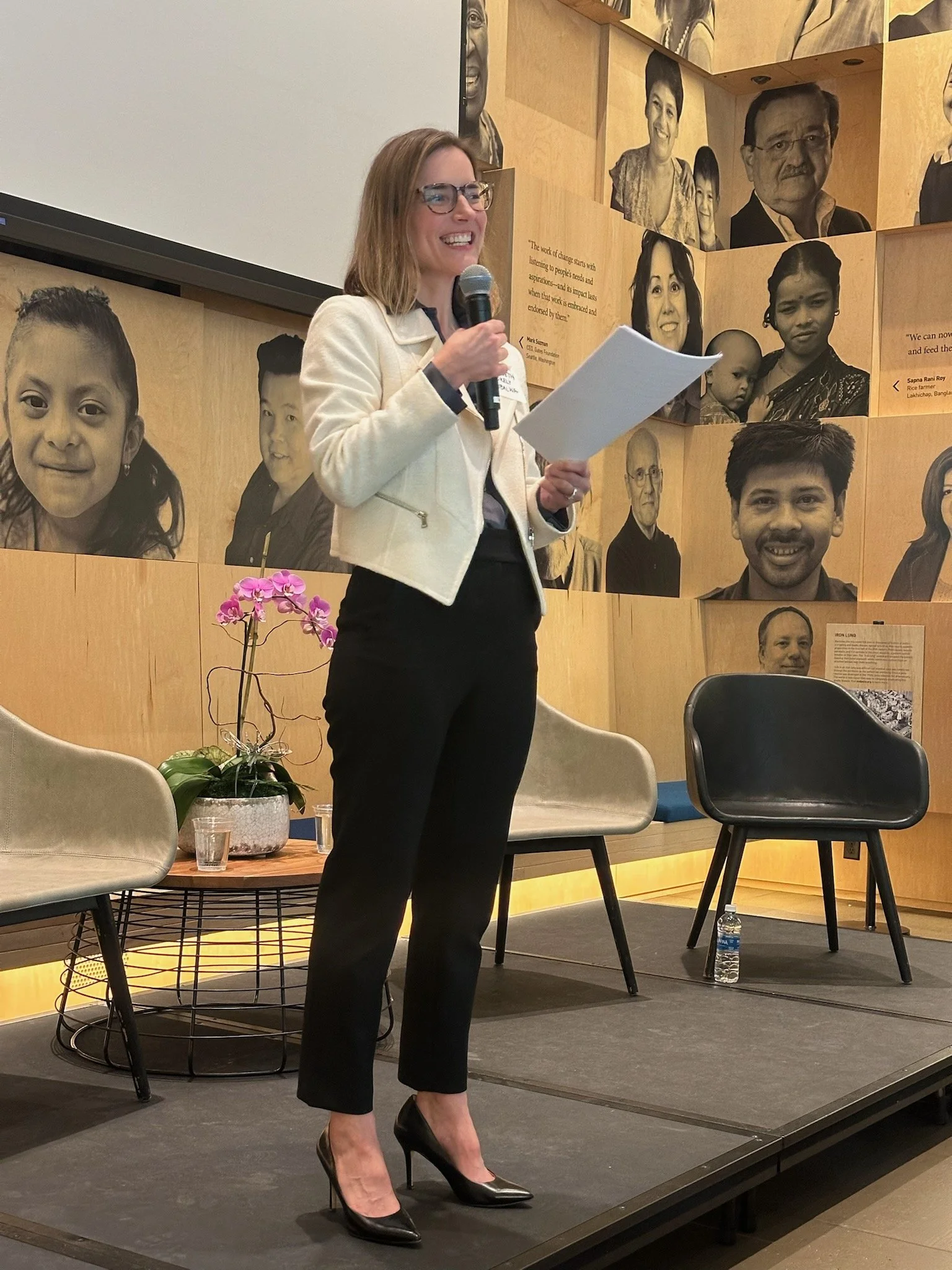 A woman in black heels and a white blazer stands on a stage holding a microphone and a piece of paper, speaking at an event. Behind her is a wooden wall decorated with black-and-white portraits of diverse individuals, with some quotes. There are chairs, a table with a potted orchid, a plastic water bottle, and a glass on stage.