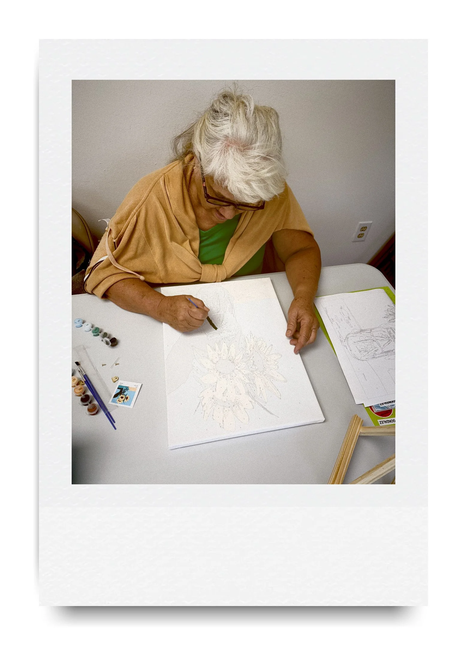 Person painting sunflower design on a canvas at a table with paint by numbers kit and brushes.