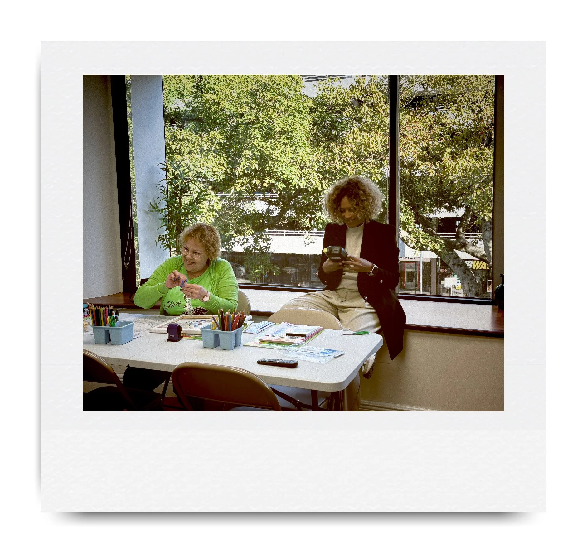 Two women sitting in a room by a large window, one at a table with art supplies and the other sitting on the window ledge, both engaged in a casual activity.
