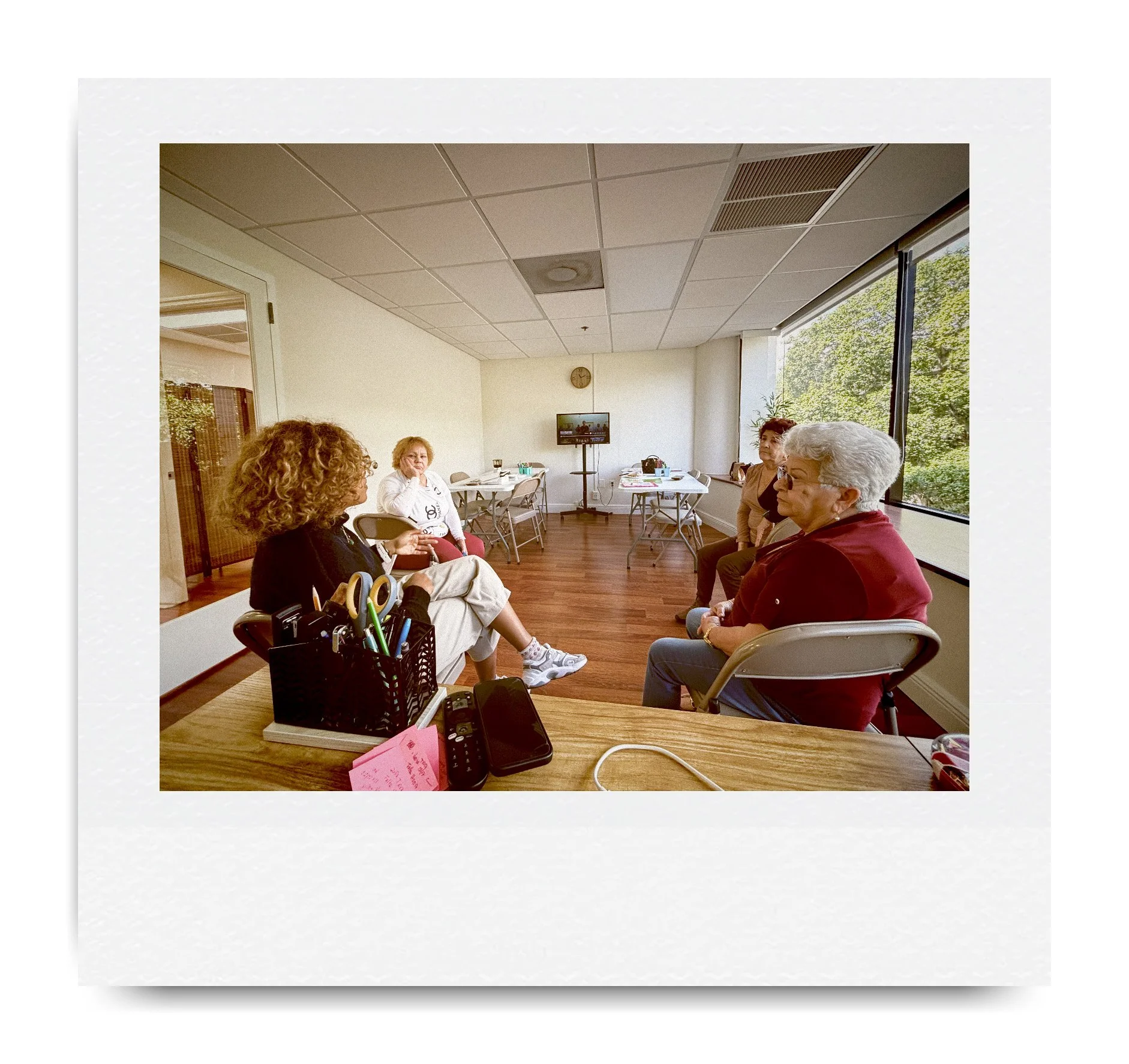A group of four people sitting in a meeting room with wooden floors and large windows. They are seated on folding chairs in a casual discussion setting. A table with office supplies and a television on a stand are visible in the background.
