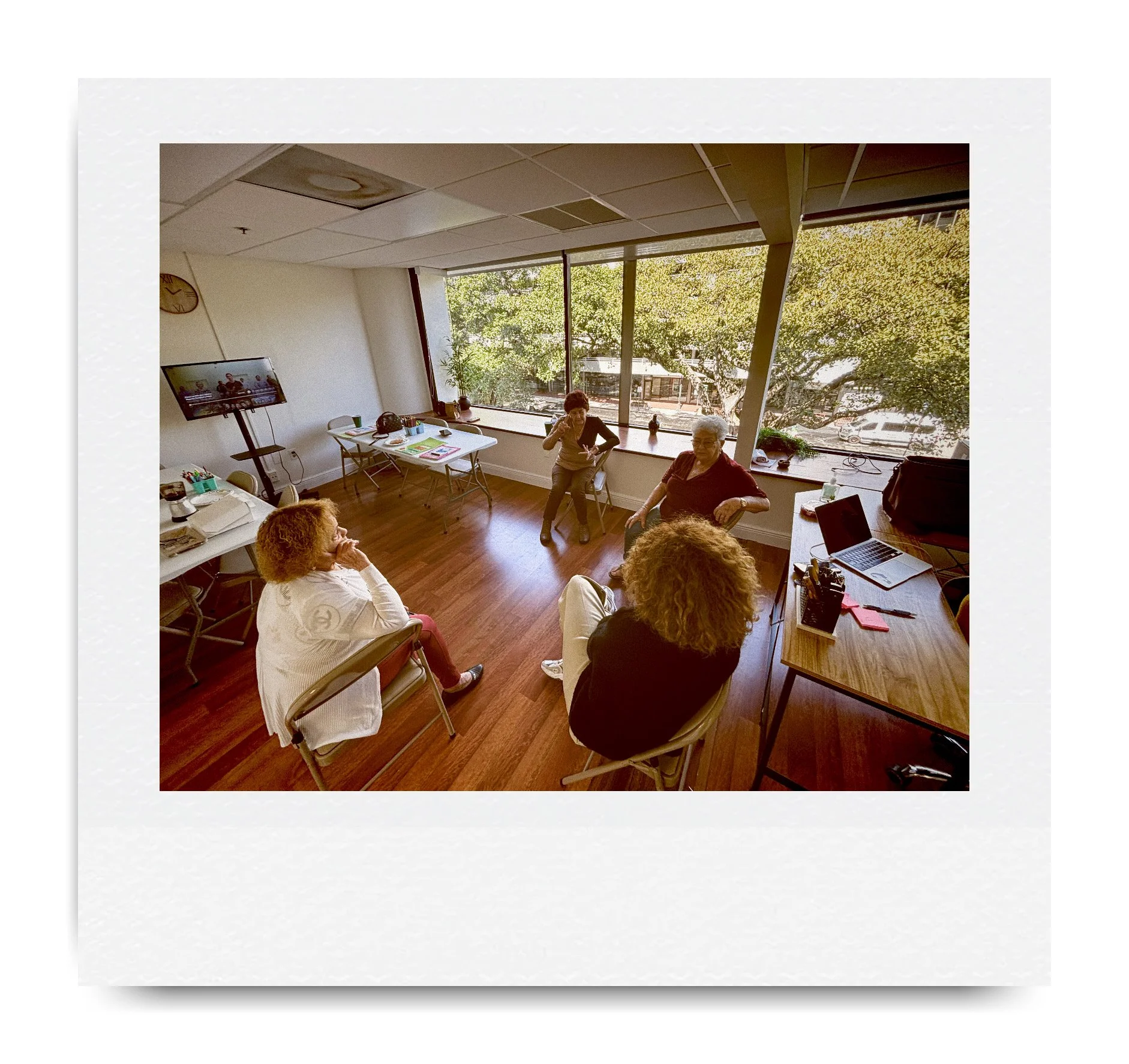 A group of people having a meeting in a well-lit office room with large window, hardwood floor, and a few tables and chairs. There is a laptop and papers on the desk. Outside the window, trees are visible. Four people are sitting in a circle.