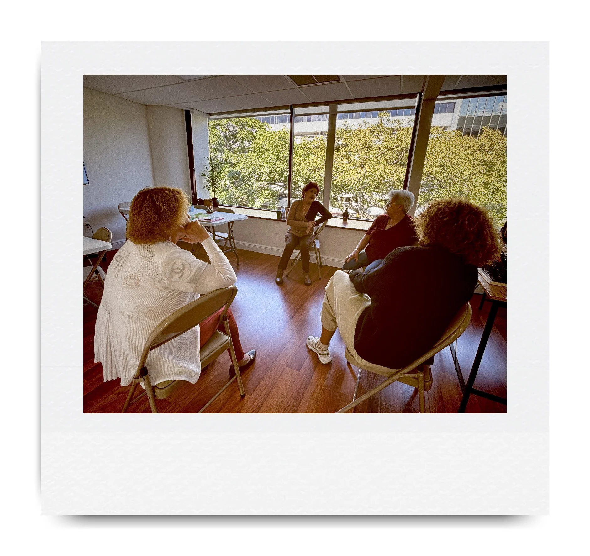 Group of people seated in a circle in a bright room with large windows and wooden floors, engaged in conversation.