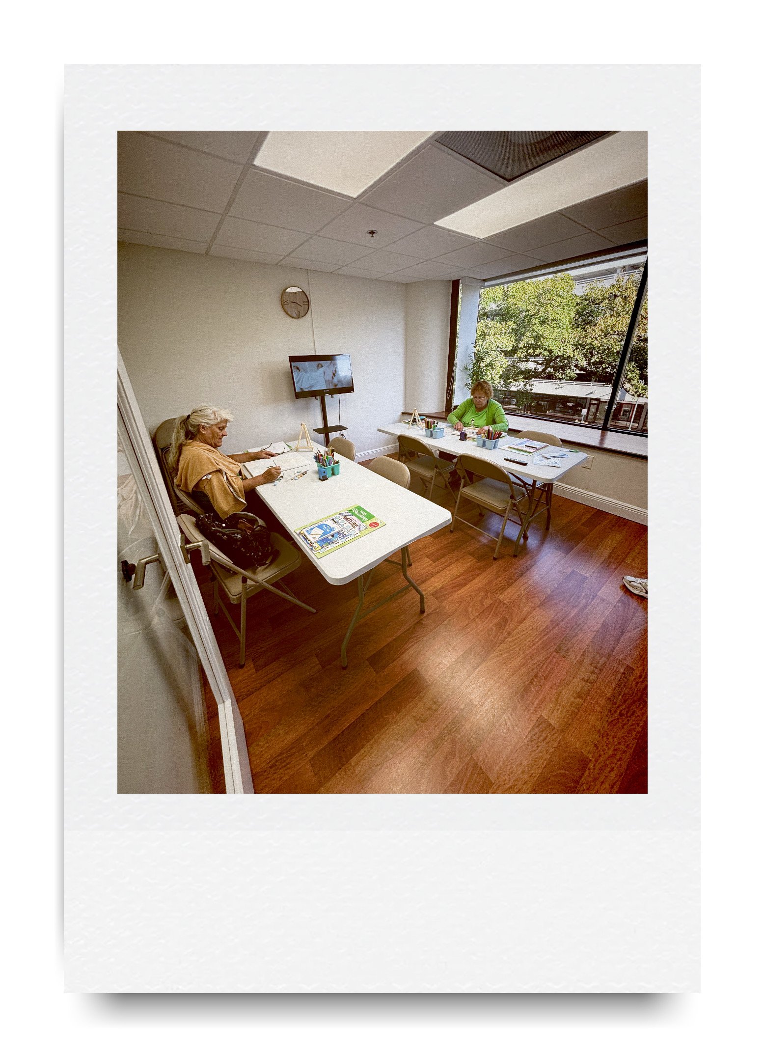 Two people sitting at tables in a classroom with art supplies and papers.