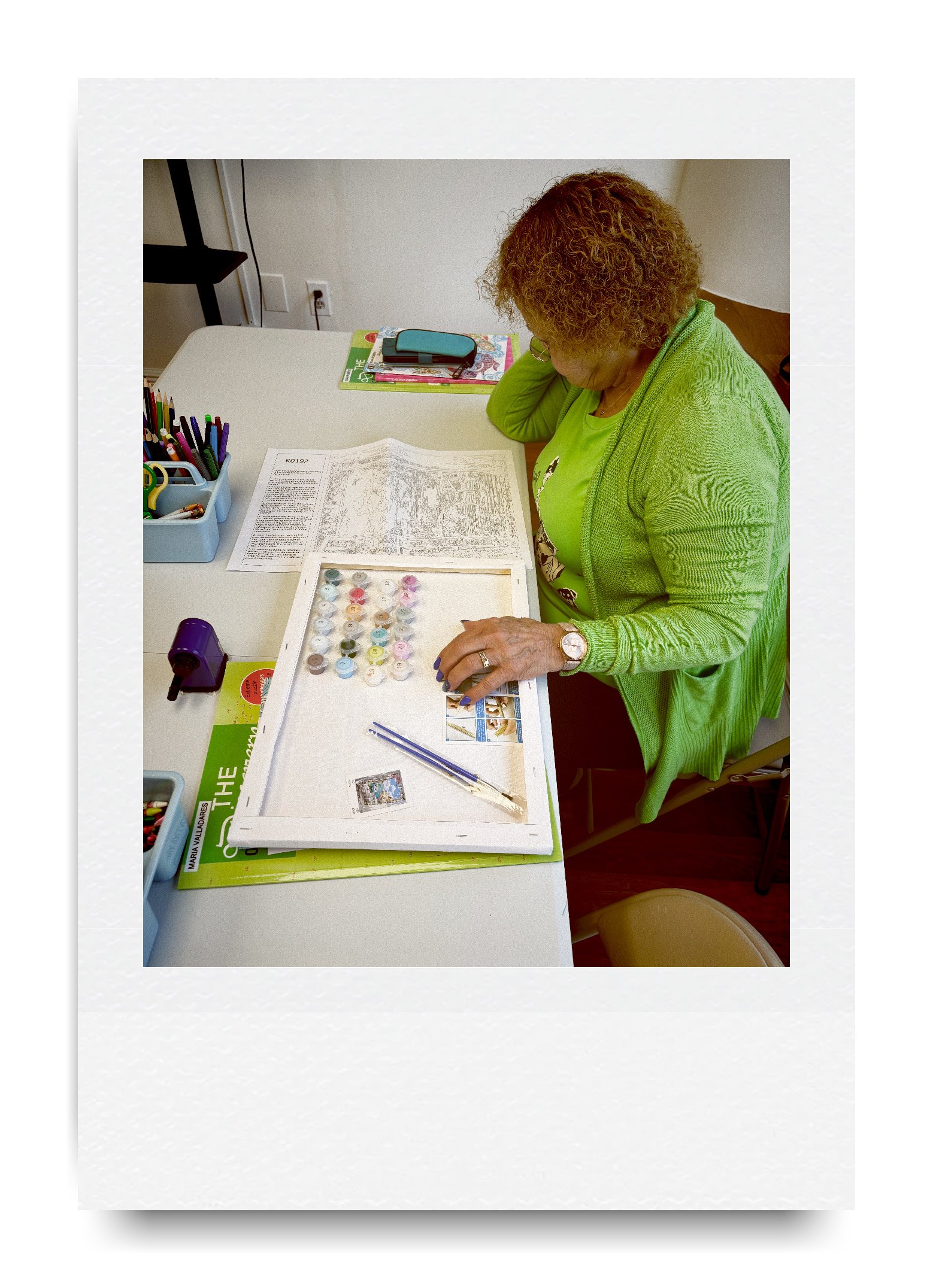 Person painting at a table with paint-by-number kit, various paint pots, colored pens, and a paper with a printed design. They are wearing a green top and sweater.