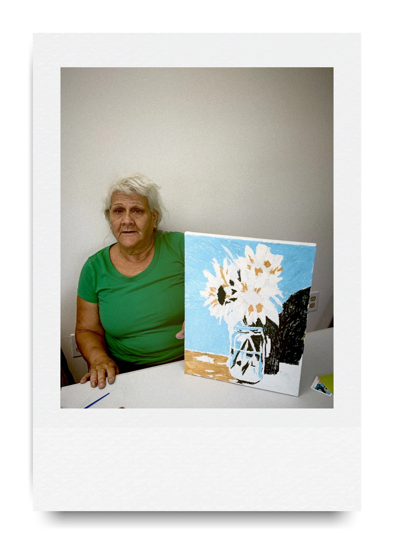 Woman in a green shirt sitting at a table with a painting of sunflowers in a jar displayed on the table.