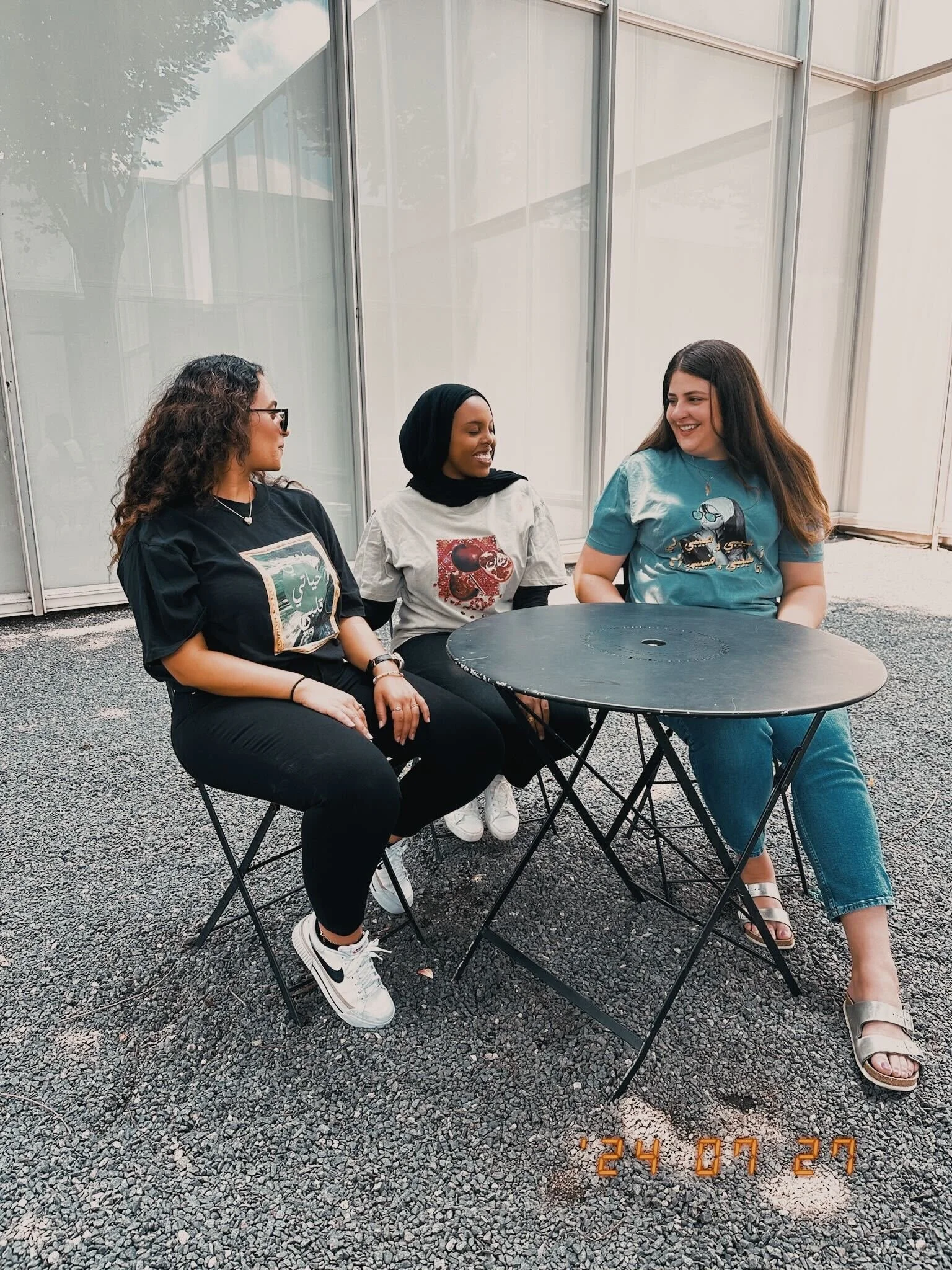 Three women sitting outside around a black table, engaging in conversation and smiling. Two are seated on black folding chairs, one woman is standing behind the chairs. All are dressed casually with graphic t-shirts. The background features glass windows and gravel ground. There is a date stamp of "24 07 27" in orange at the bottom of the image.