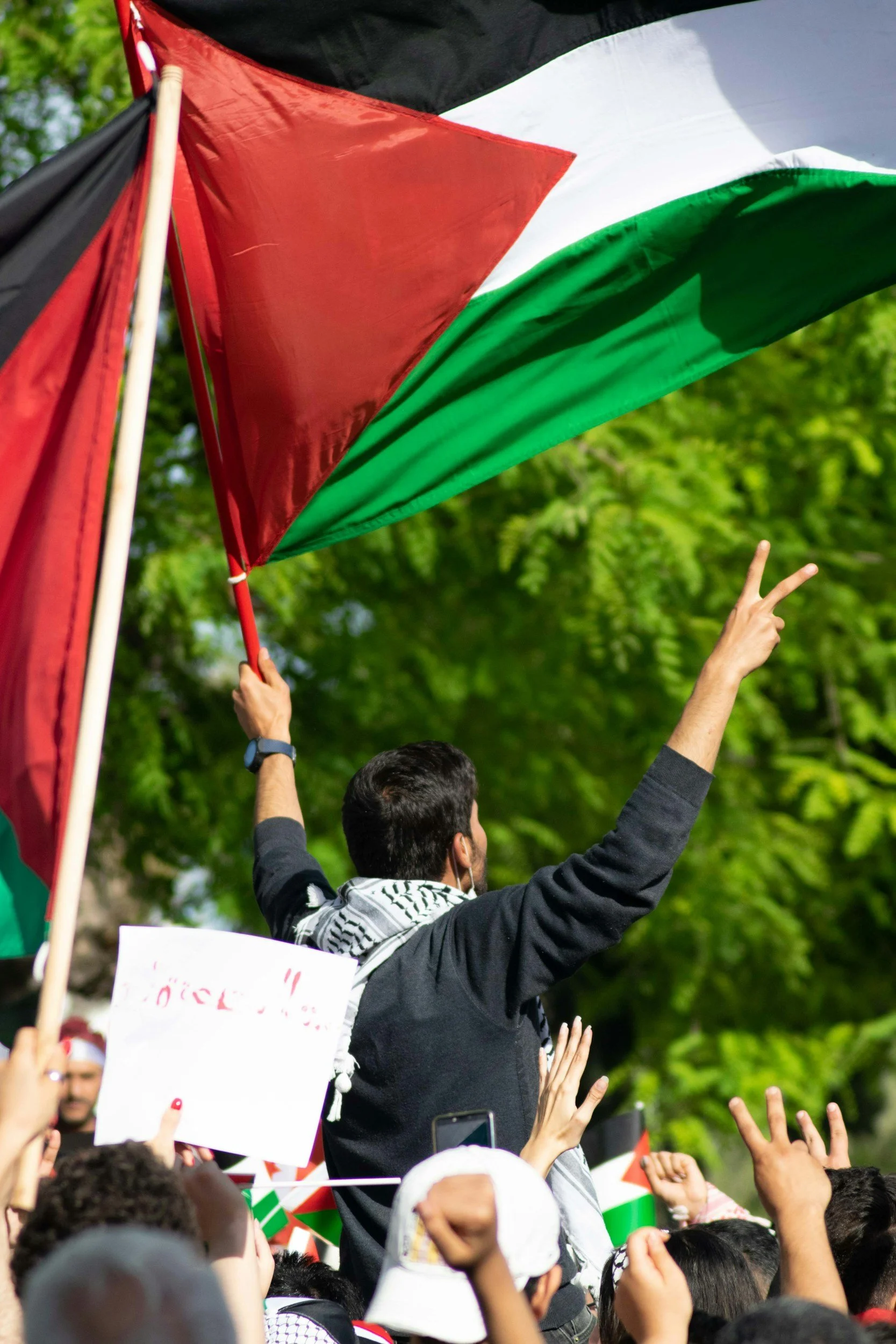 A man holding a Palestinian flag and making a peace sign with his fingers amidst a crowd of people during a demonstration.