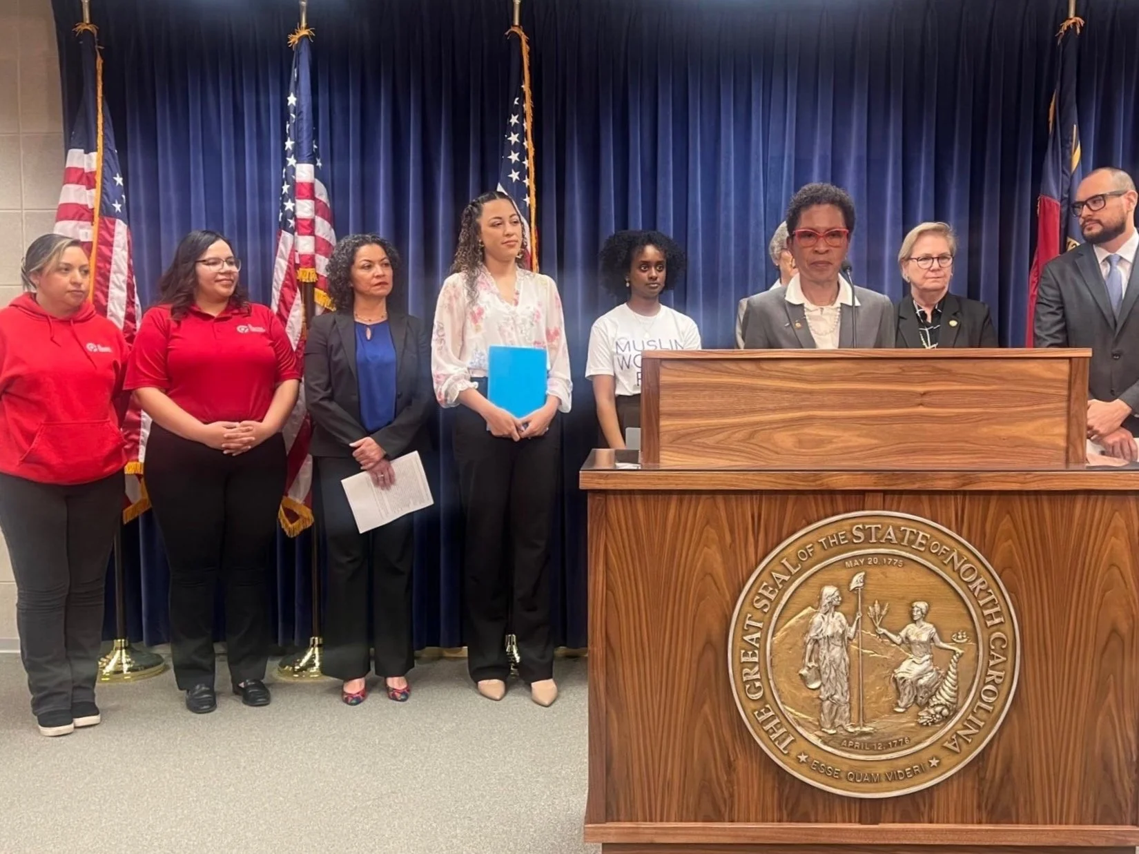 Group of diverse women and men standing behind a wooden podium with a seal of North Carolina, in front of a blue curtain with American flags, in a formal setting.
