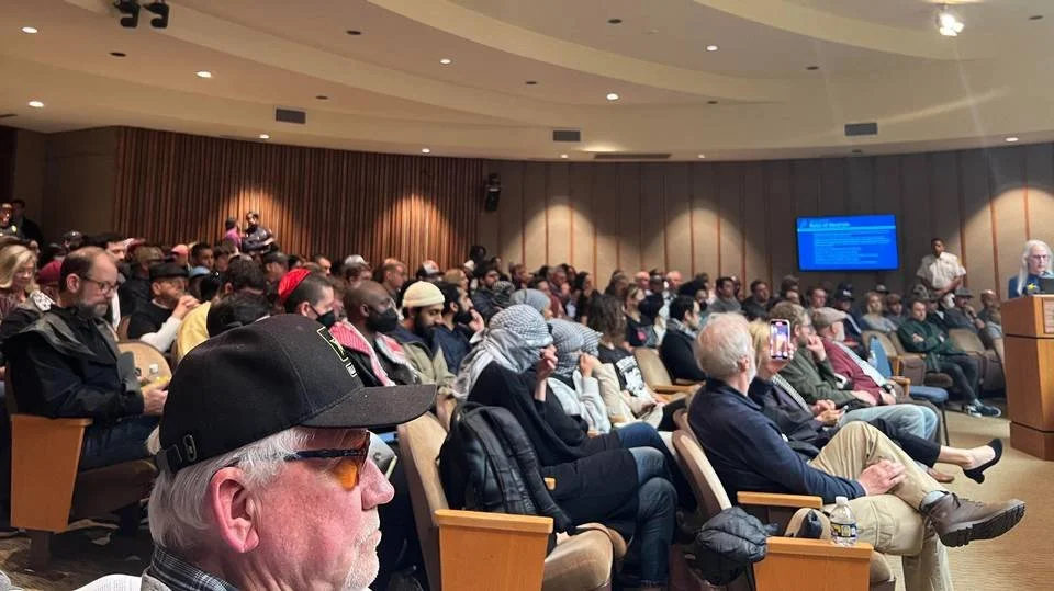 Large group of people attending a conference or seminar in an auditorium, listening to a speaker at a podium on the right, with some audience members taking photos or notes.