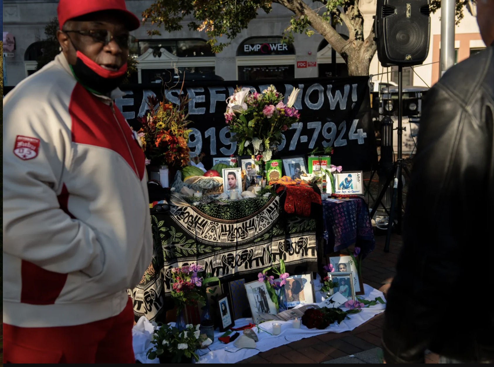 Memorial altar with flowers, photos, candles, and personal items for an individual, set up outdoors with people nearby.