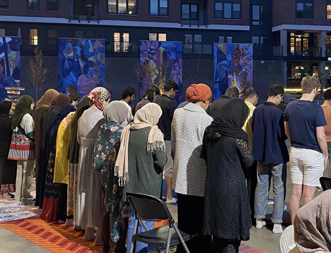 A diverse group of people standing and praying in line outdoors near a building decorated with colorful murals at dusk.