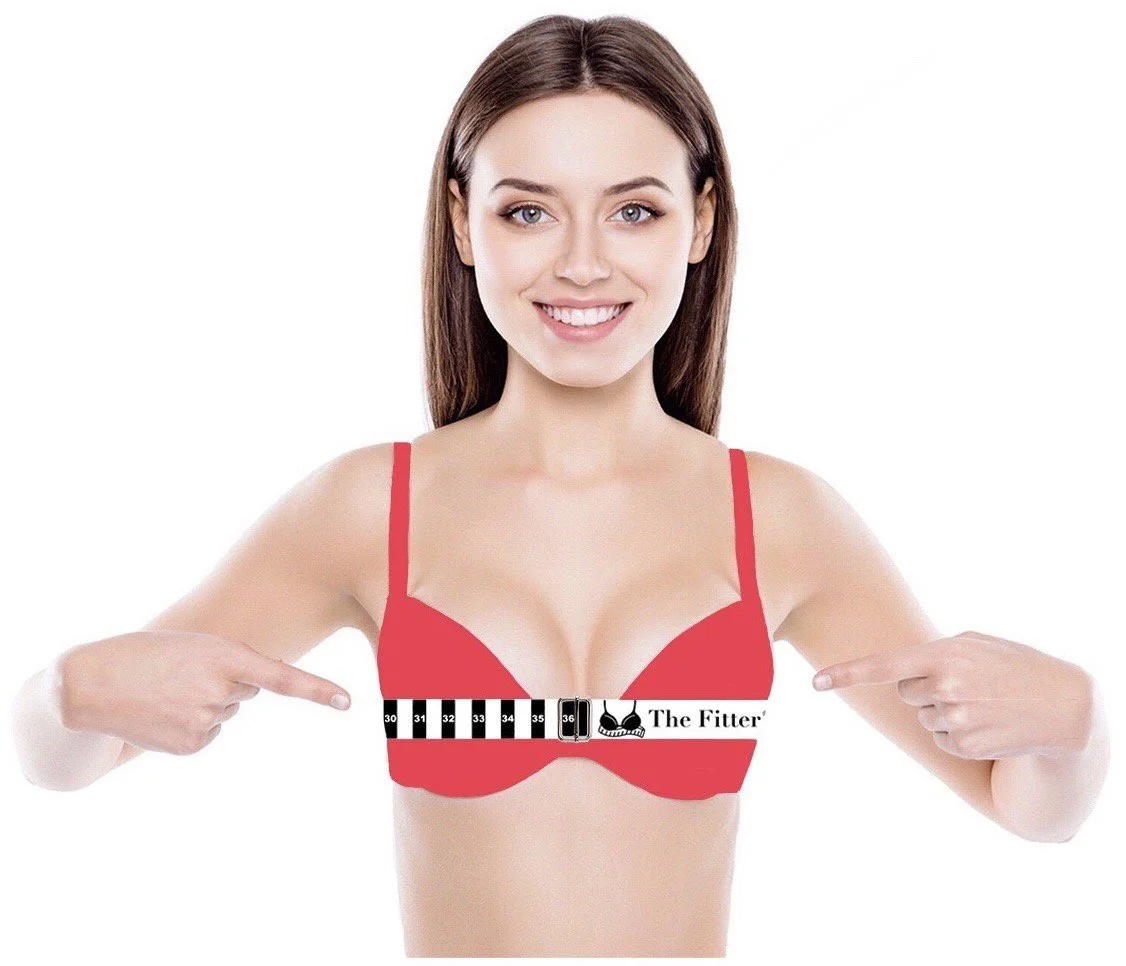 A woman with long brown hair smiling, pointing to her red bra that has a measuring tape labeled 'The Fitter' wrapped around it.