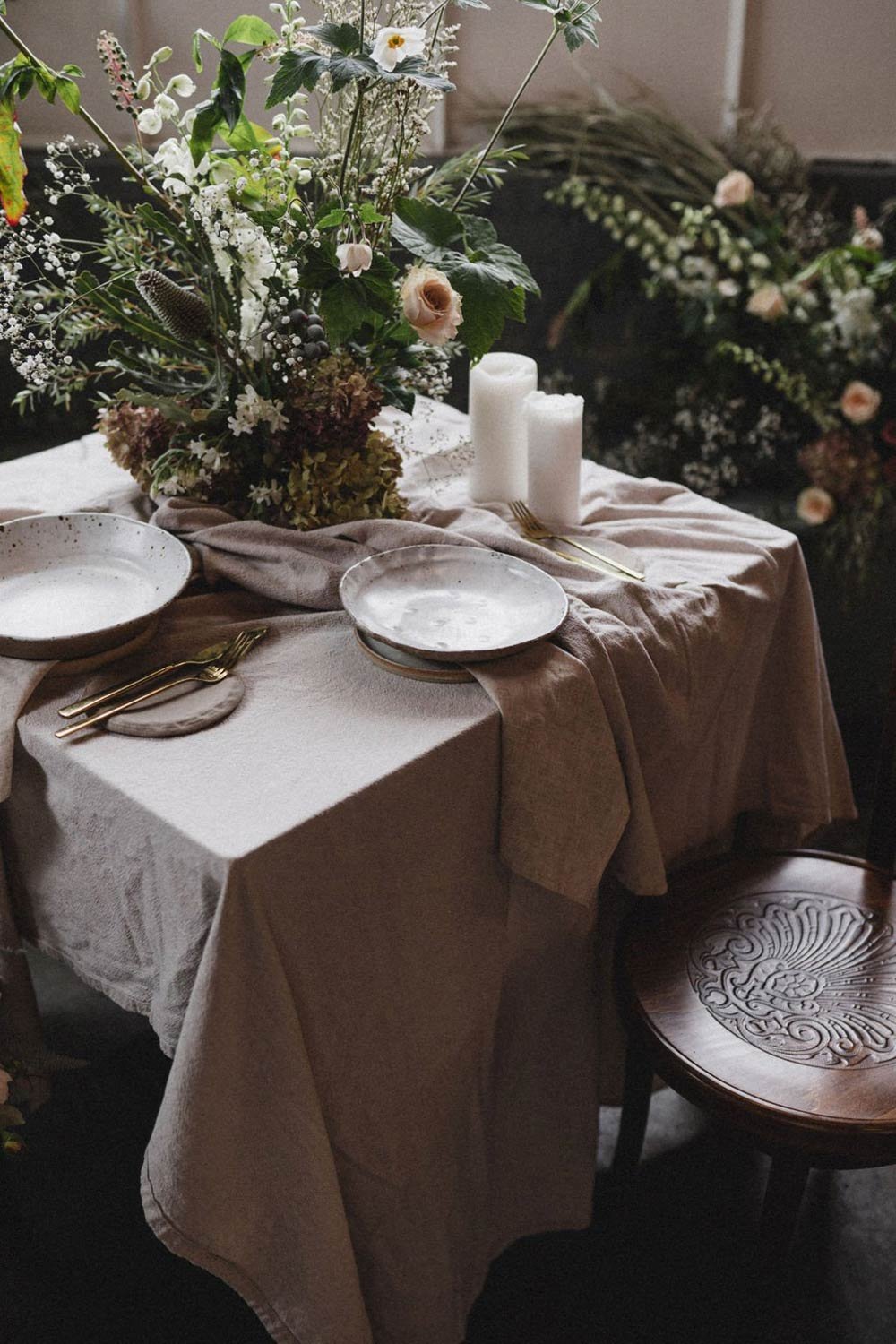 A table set for a dinner with a floral centerpiece, two candles, gold cutlery, and a beige tablecloth, with pink roses and greenery in the background. Avenue Studio Co. Surrey Hills. Melbourne. Wedding in style.