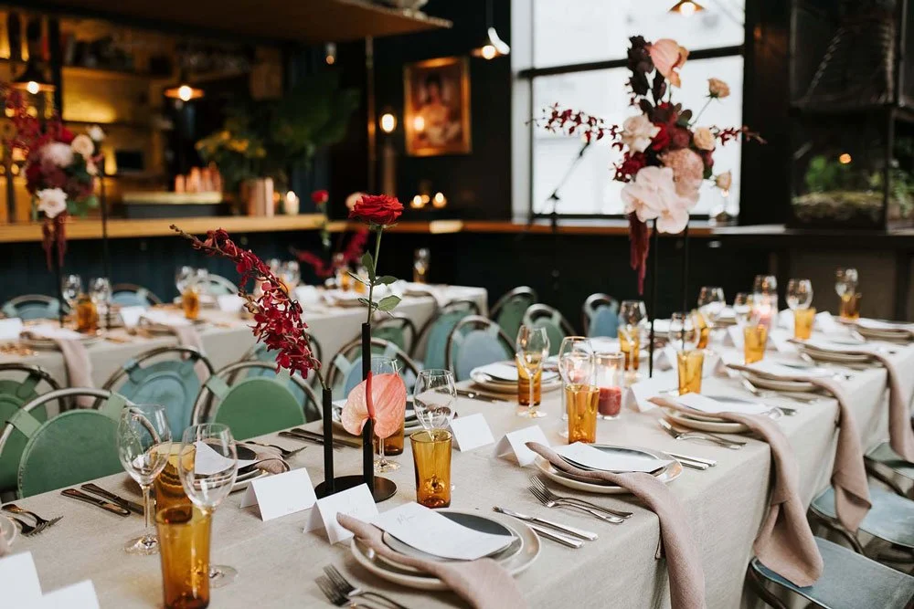 Elegant restaurant dining table set with floral centerpieces, wine glasses, water glasses, white plates, cutlery, and beige napkins in a well-lit room. Avenue Studio Co. Surrey Hills. Melbourne. Wedding in style.