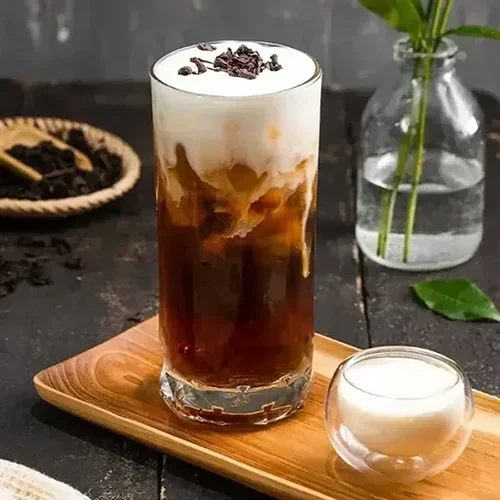 Iced coffee with whipped cream and chocolate shavings in a tall glass, served on a wooden tray, with a small bowl of cream or milk nearby, and a glass jar with green plant in the background.