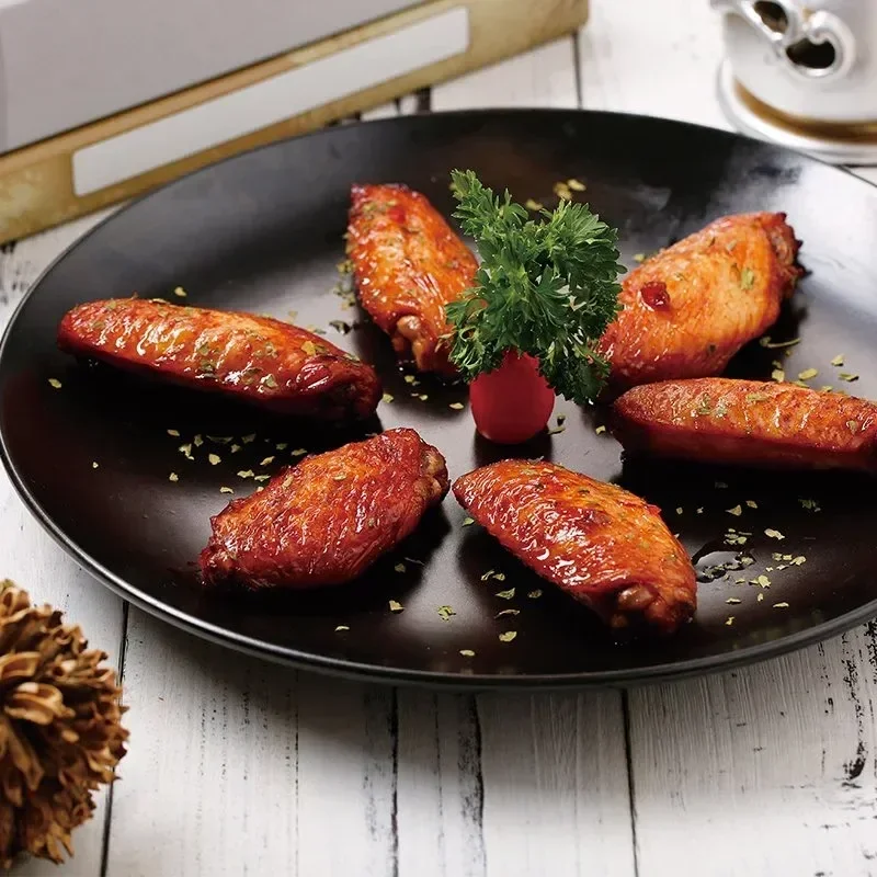 A black plate with six glazed chicken wings garnished with parsley and a radish, on a white wooden surface.