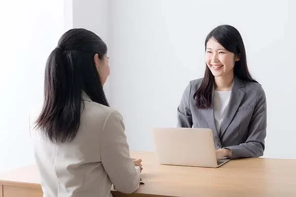 Two women sitting at a table having a conversation, one has a laptop in front of her, in a bright, minimal office space.