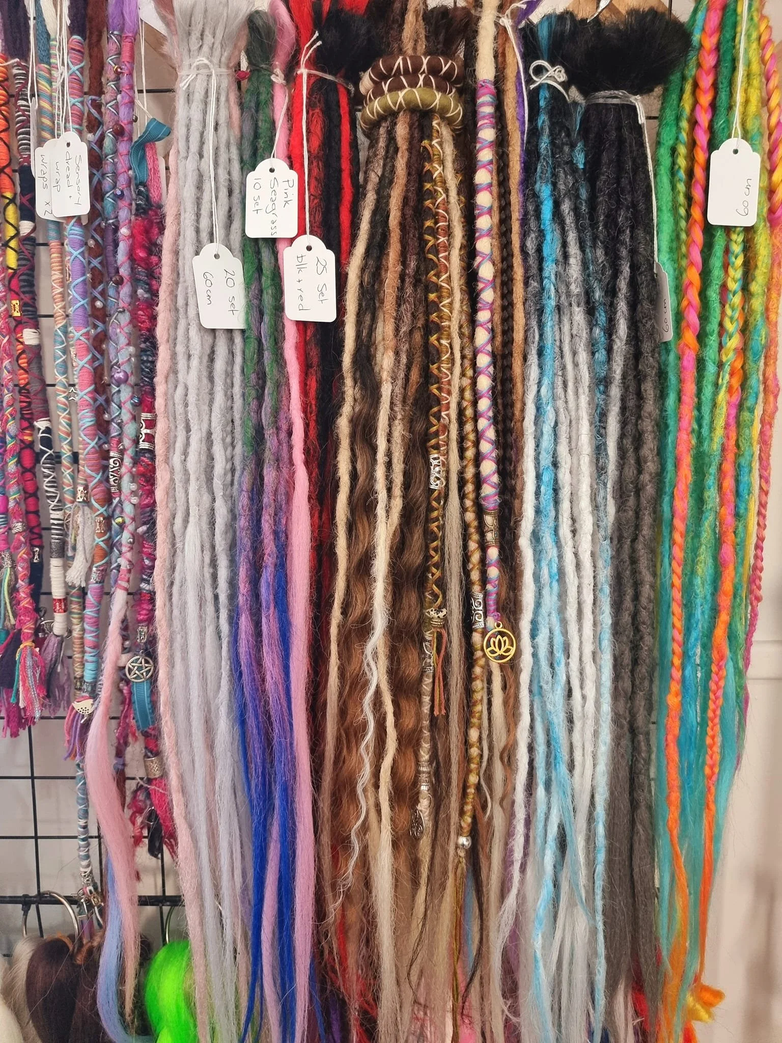 Colorful display of various dreadlock extensions and hair accessories hanging on a metal grid wall, with labels indicating different styles, lengths, and colors.
