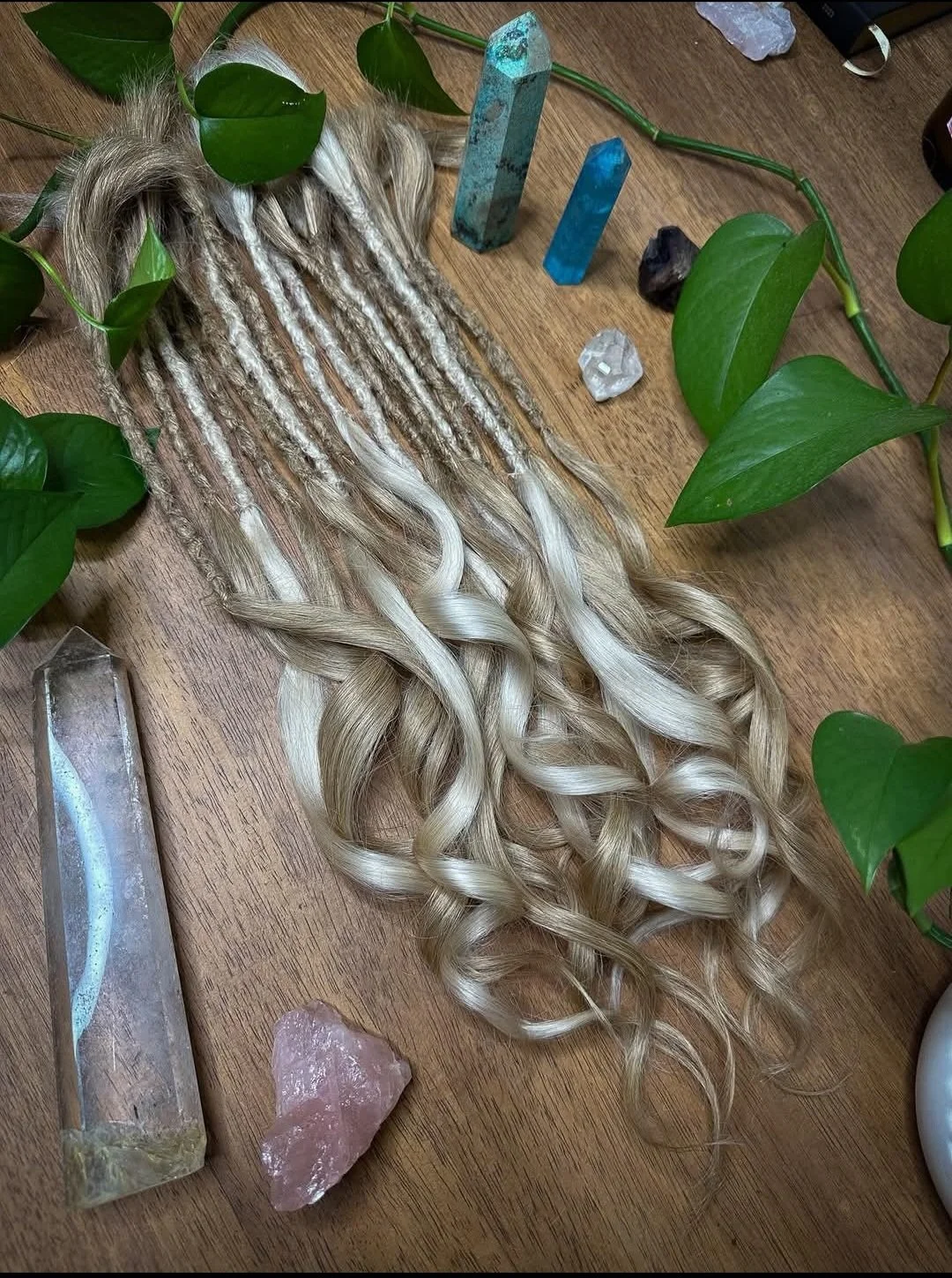 A bundle of blonde, wavy synthetic or natural hair extensions on a wooden surface, surrounded by green leafy vines and various crystals and gemstones, including quartz, rose quartz, and two large mineral towers.