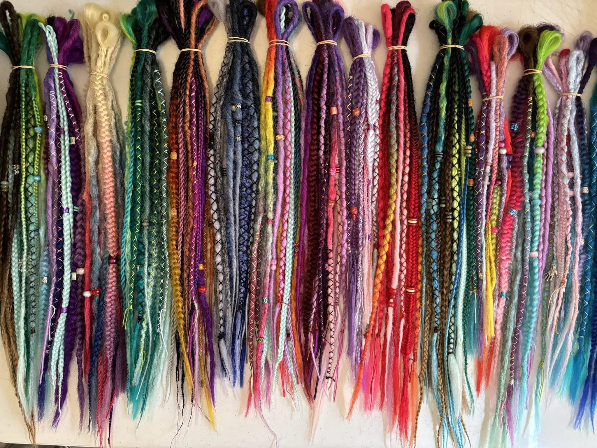 Colorful yarn braids hanging on a wall, arranged in a row with some beads and accessories on some of the braids.