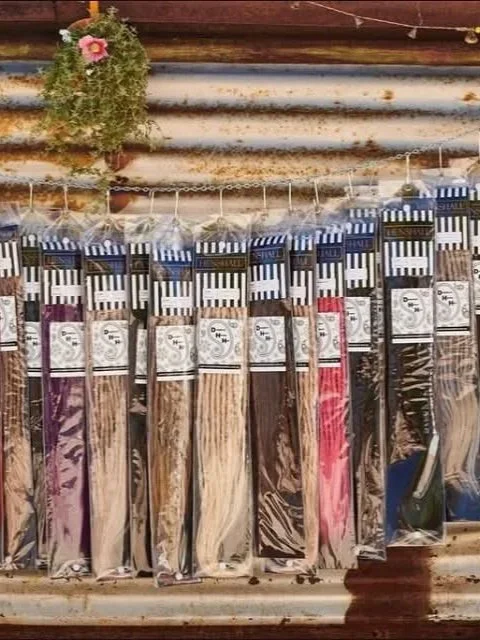 Display of synthetic hair extensions and hairpiece accessories hanging against a rustic, rusty metal wall. The setup includes various braided and straight hairpieces, some with colorful, faux-dreadlock styles, and decorative beads and hooks.